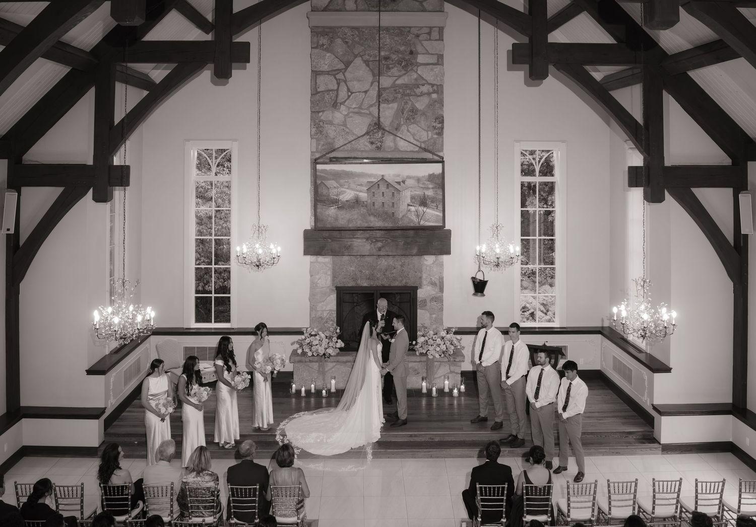 Black and white photo of an elegant wedding ceremony inside a chapel with vaulted ceilings, chandeliers, and a stone fireplace.