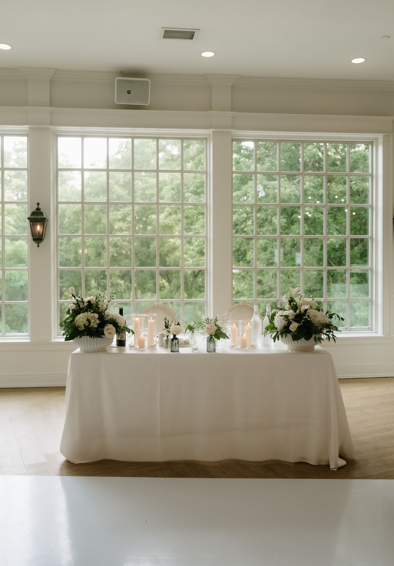 Elegant sweetheart table with white florals and candles set before large windows overlooking a garden.