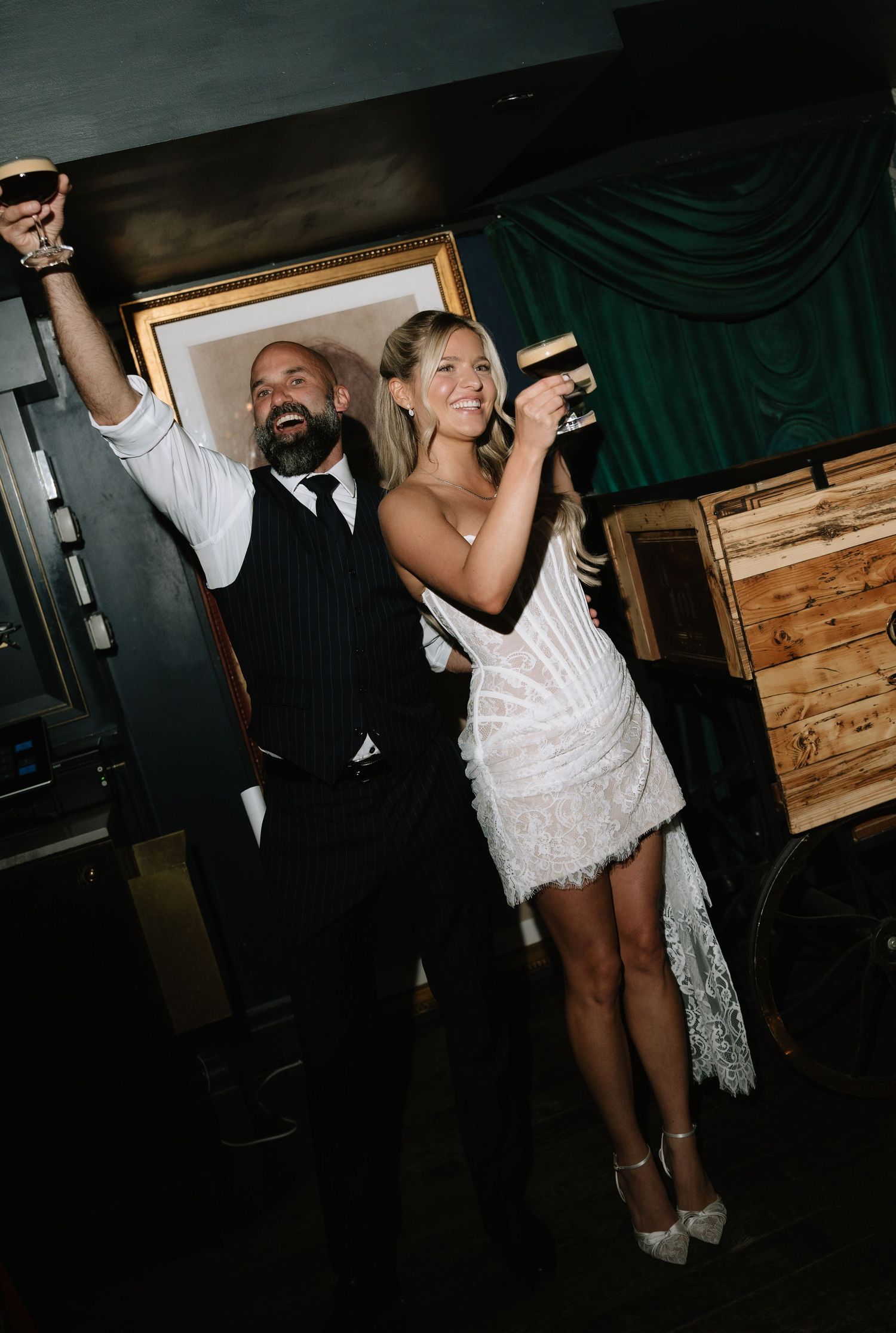 A joyful couple in wedding attire raises glasses in celebration at a dimly lit reception venue.