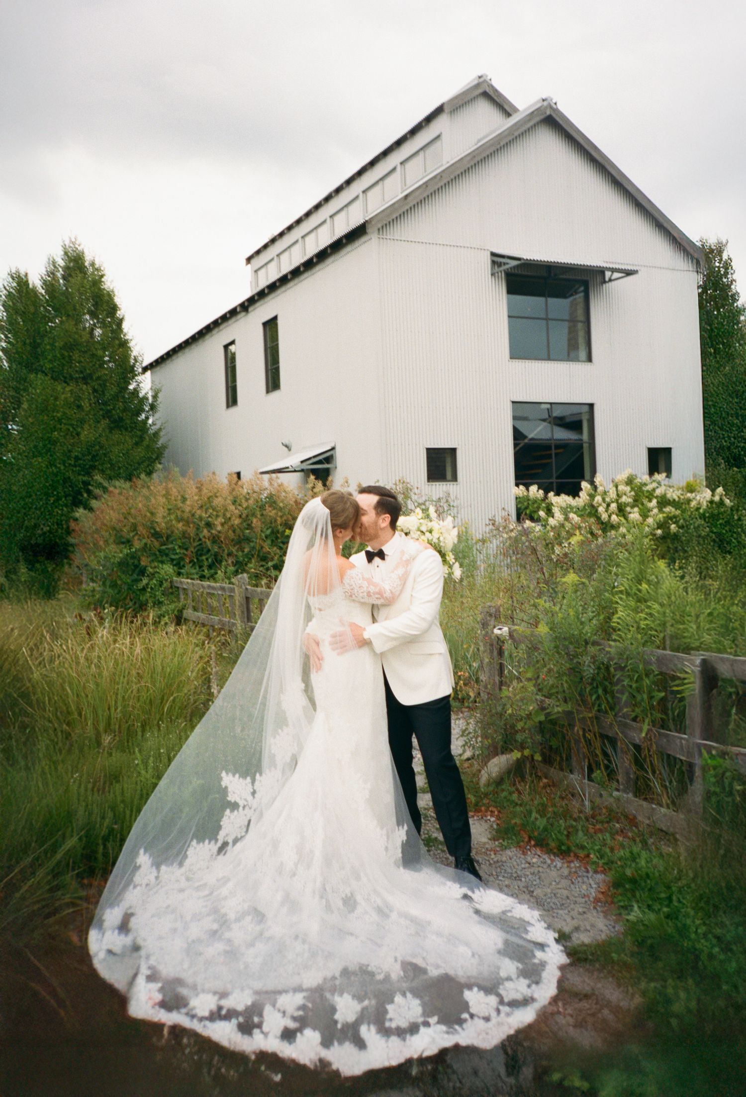 Bride and groom share a kiss outside white modern barn venue, cathedral lace veil flowing in the breeze.
