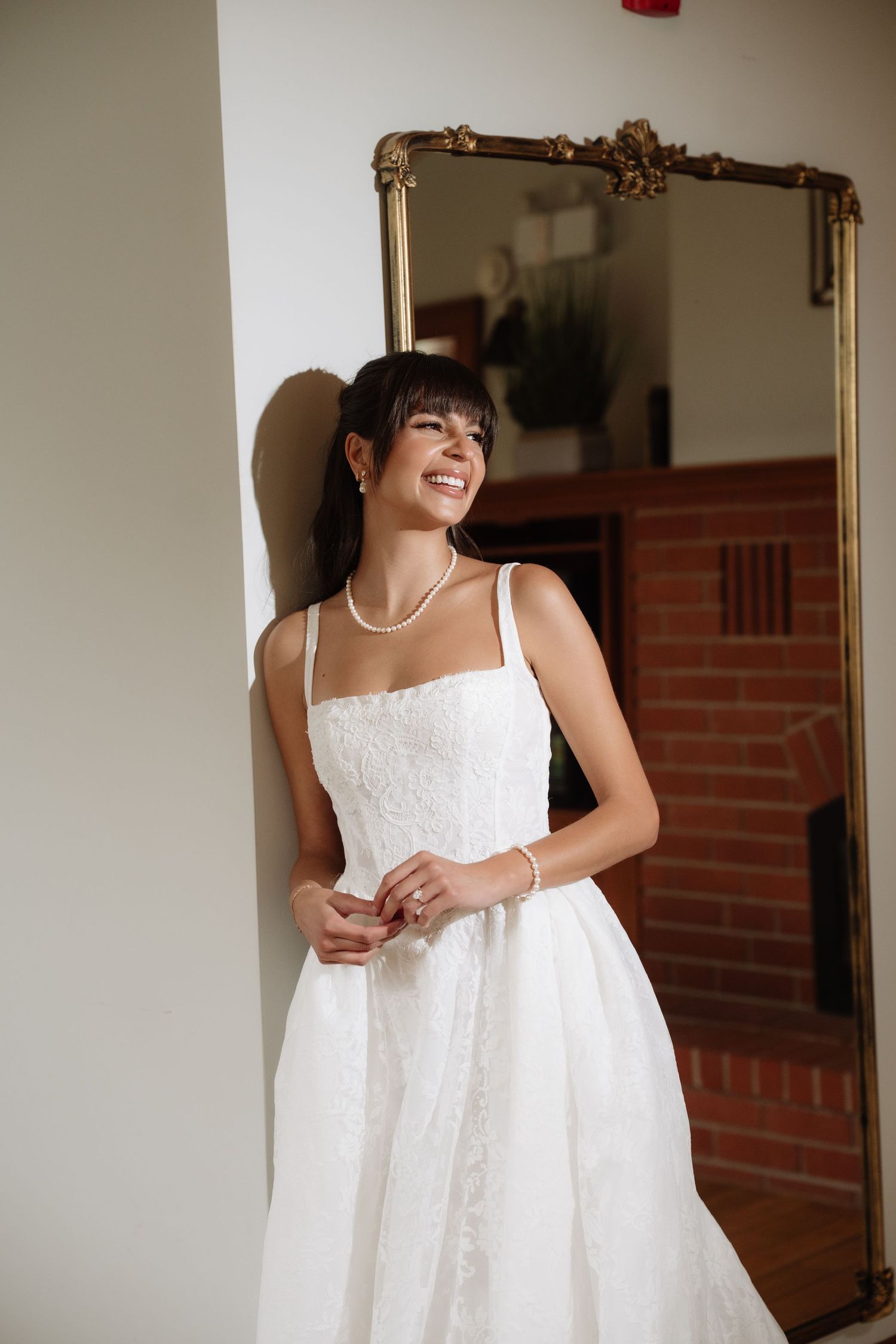 Smiling bride in white lace A-line wedding gown with pearl necklace, leaning against wall near gold mirror.