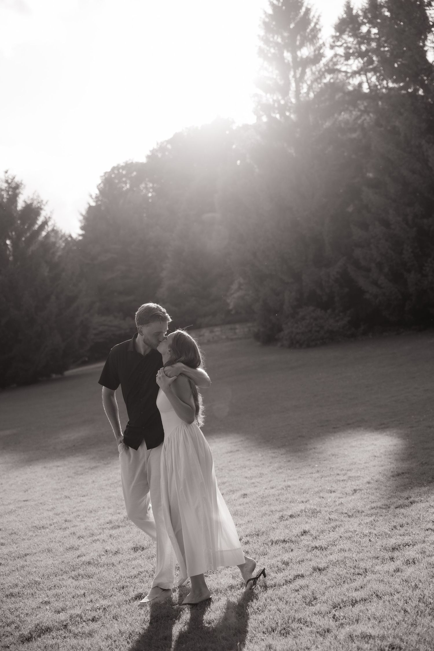 Black and white photo of a couple sharing a romantic moment in a sunlit garden with tall trees.