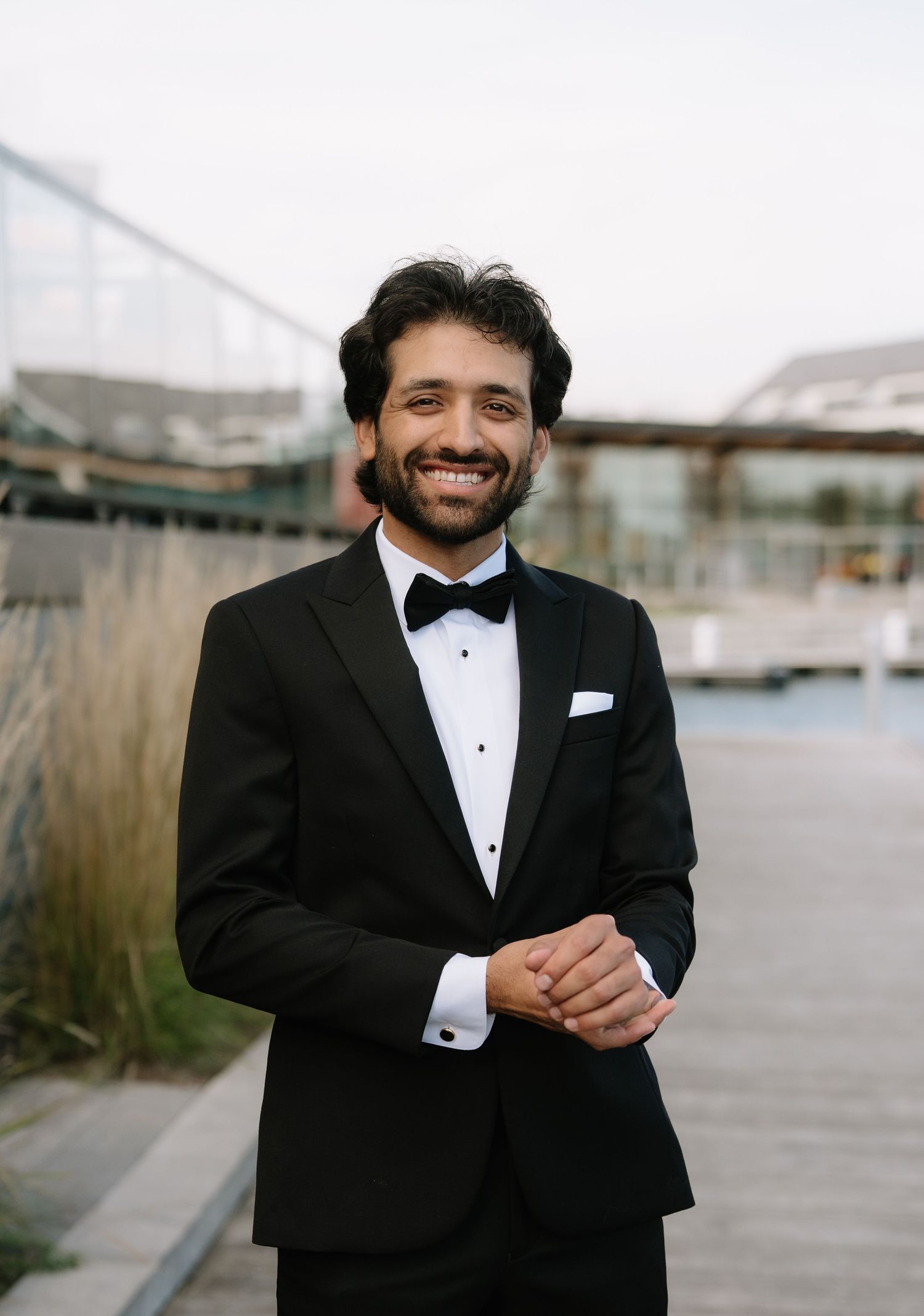 Smiling groom in black tuxedo with bow tie and white pocket square poses outdoors near waterfront.