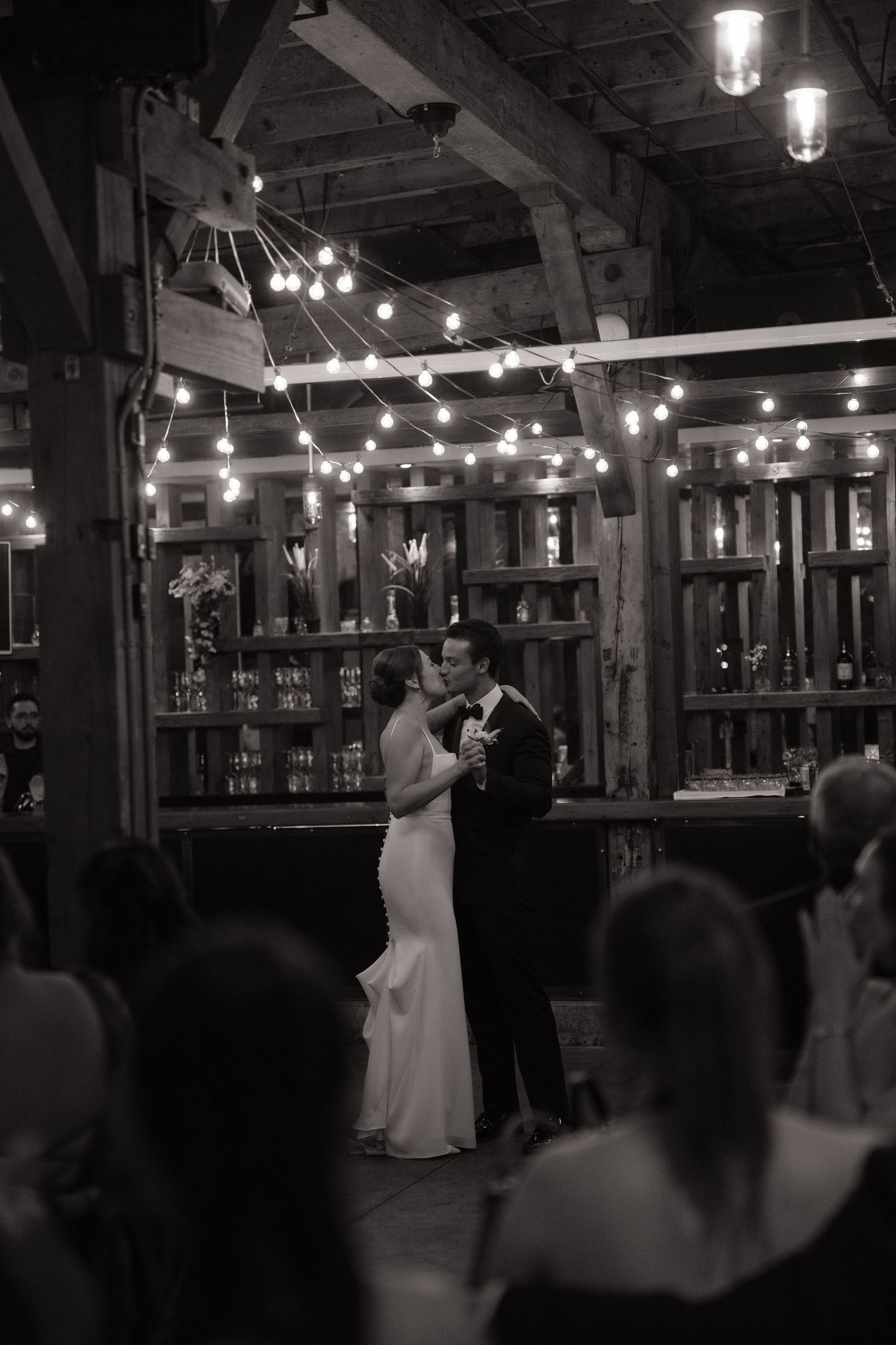 Black and white photo of bride and groom sharing first dance under string lights in a rustic barn wedding venue.