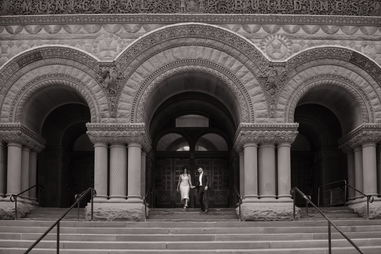 Black and white photo of newlyweds posing beneath ornate arched entrance of a historic stone building.