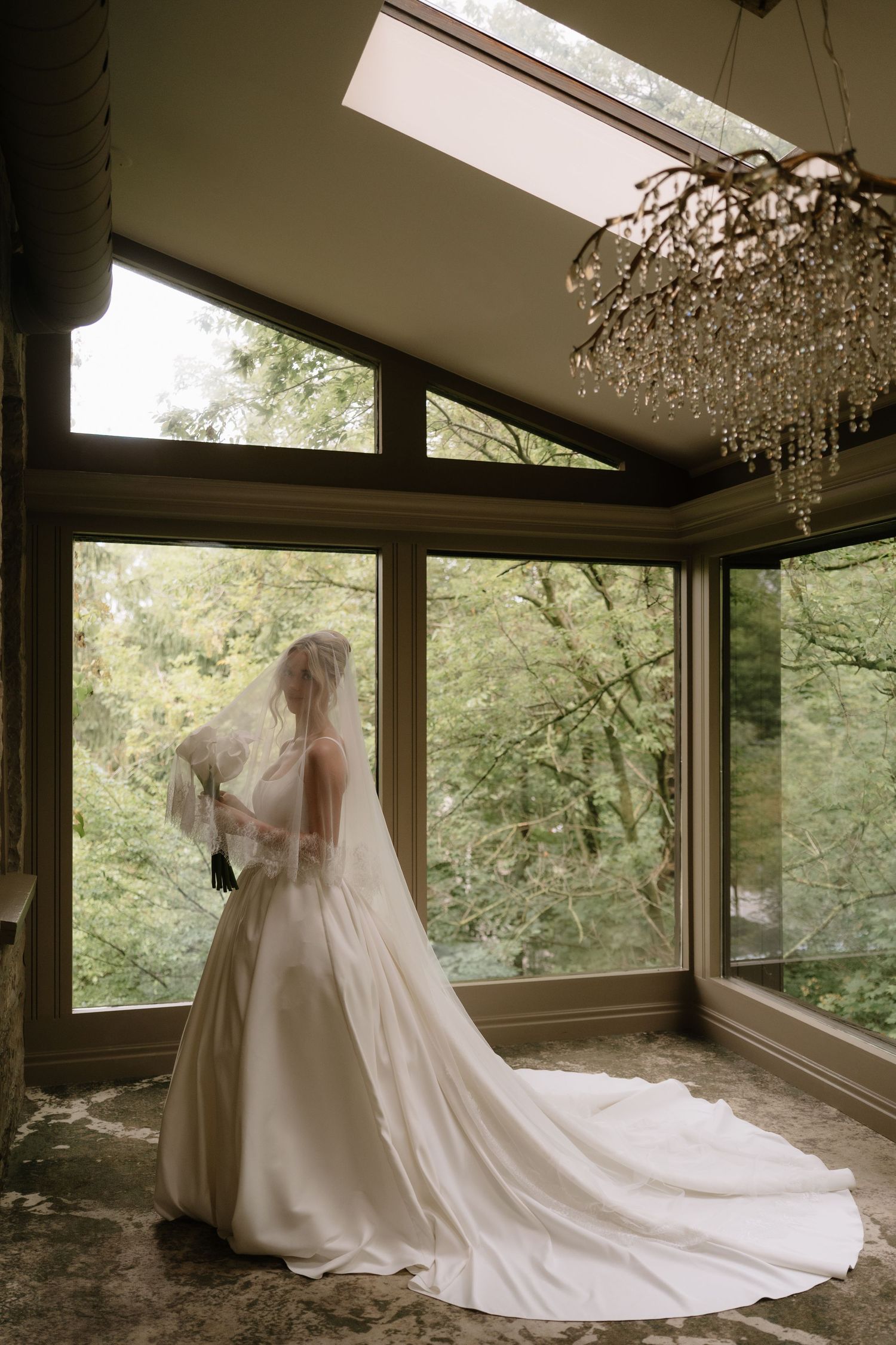 Bride in a white gown and veil stands by large windows with a crystal chandelier overhead, lush greenery visible outside.