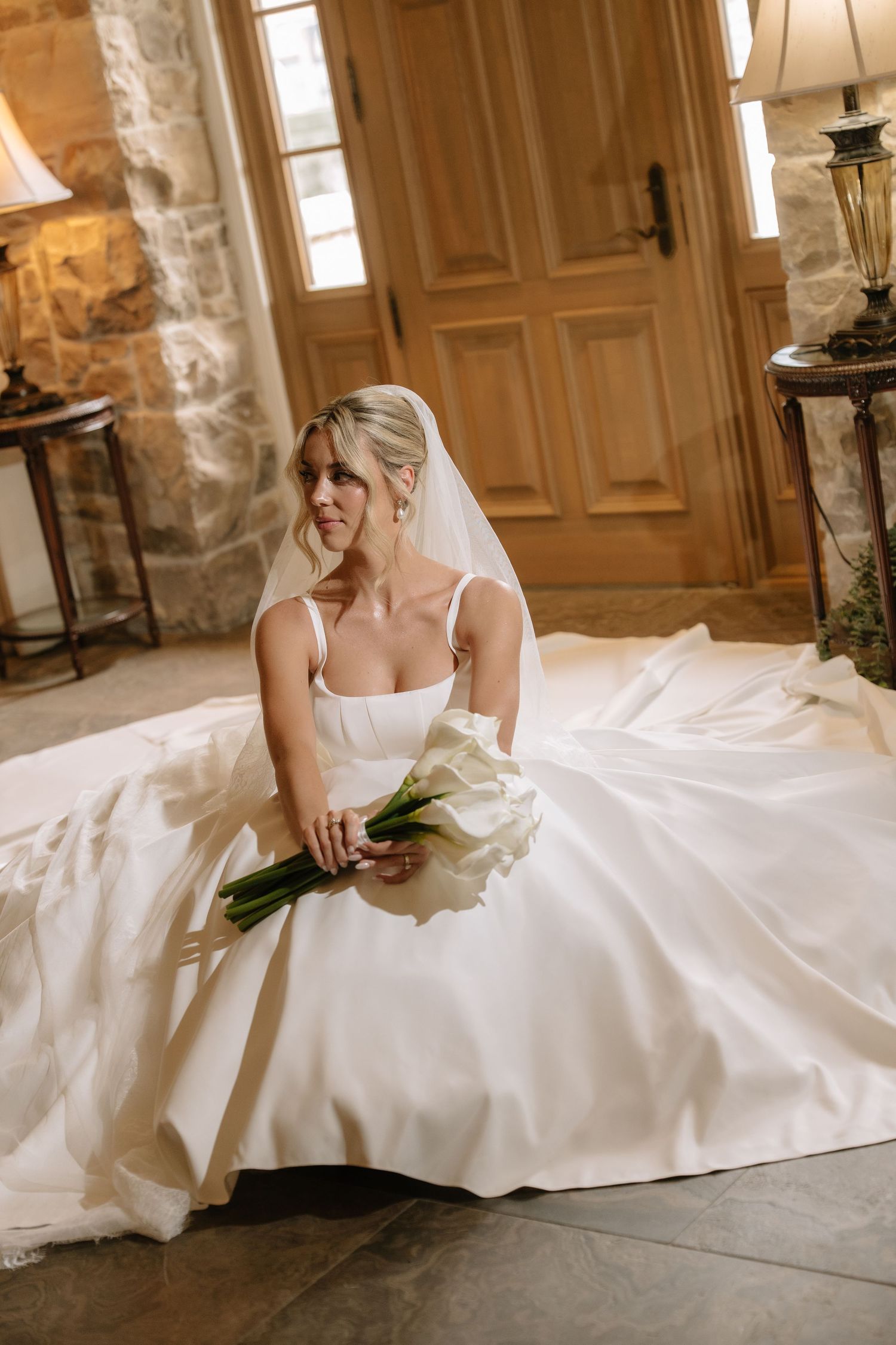 Bride in white ball gown with veil sits on stone floor holding calla lily bouquet near wooden doors.