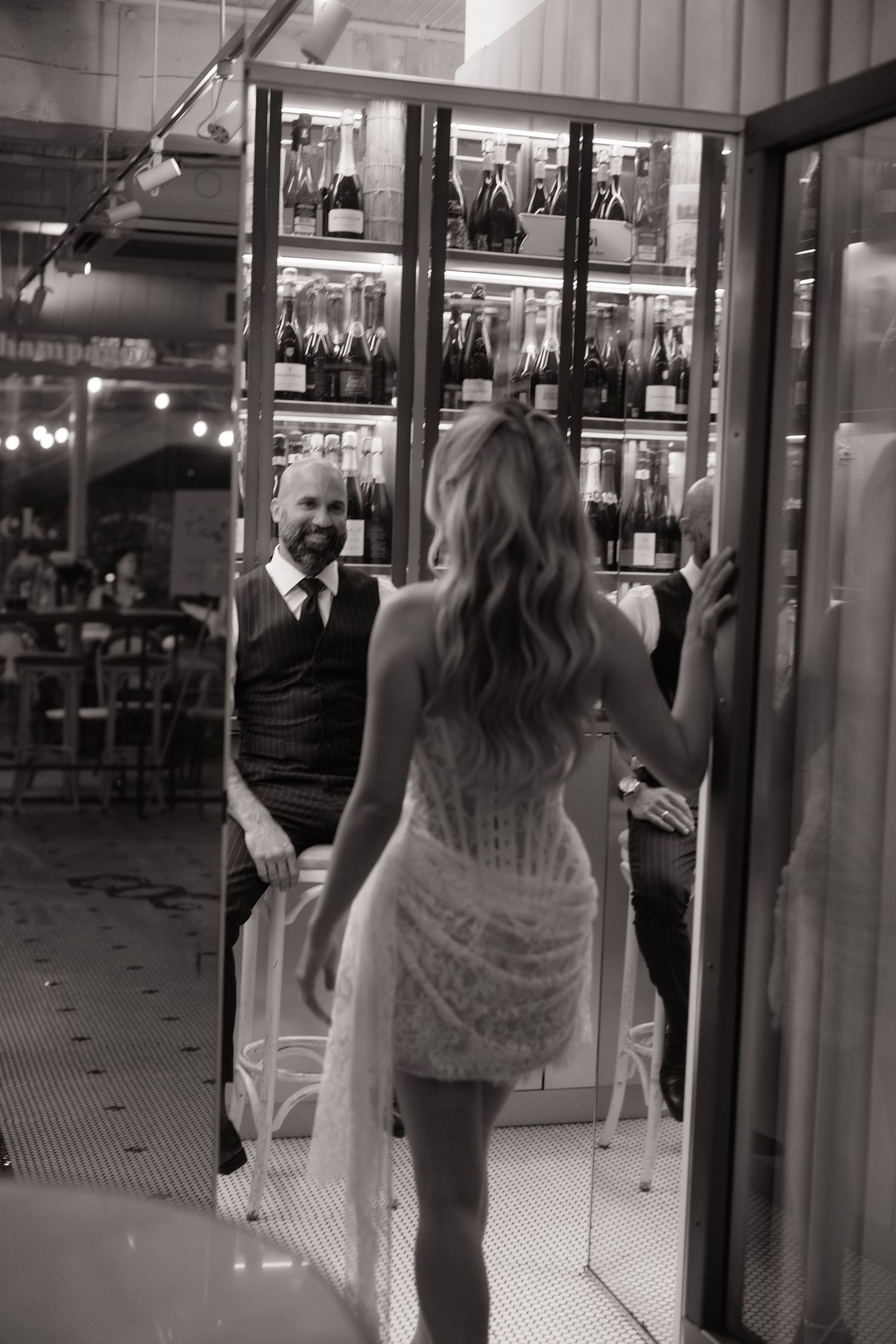 Black and white photo of a bride in a short lace dress approaching a smiling groom seated near a wine display.