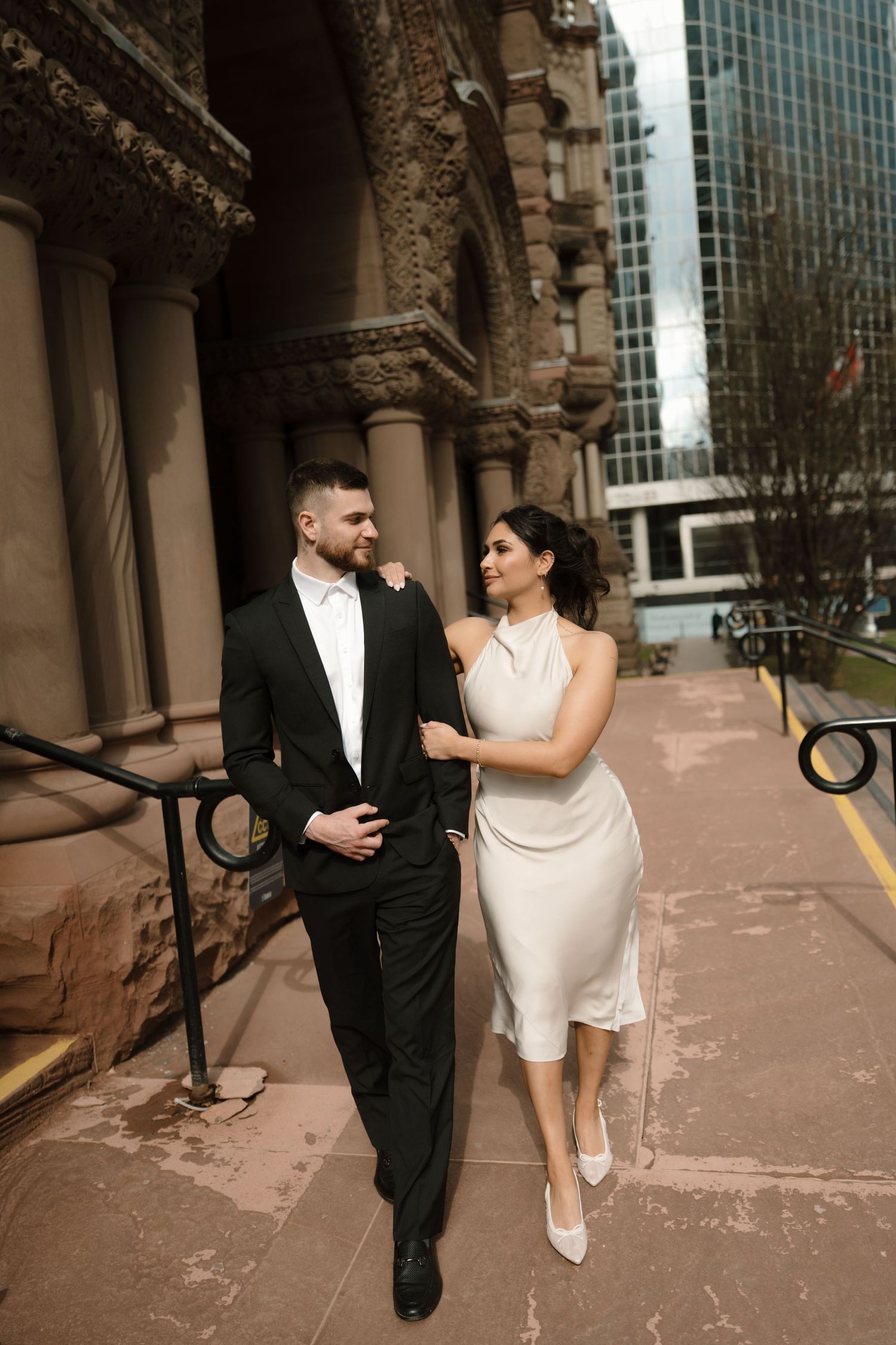 Couple in wedding attire share a romantic moment outside historic stone building in urban setting.