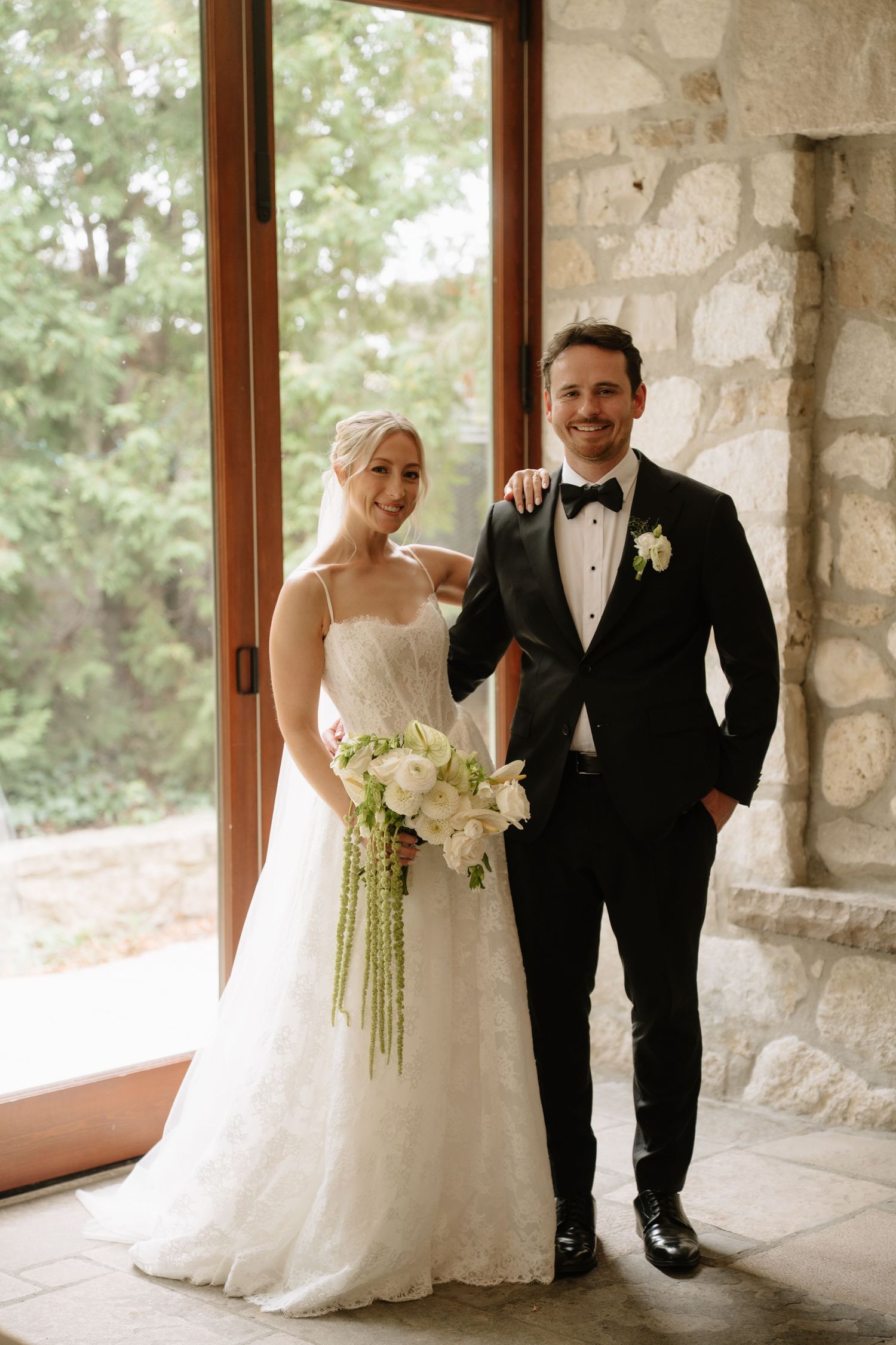 Bride in white lace gown holding floral bouquet stands with groom in black tuxedo near stone wall and glass doors.
