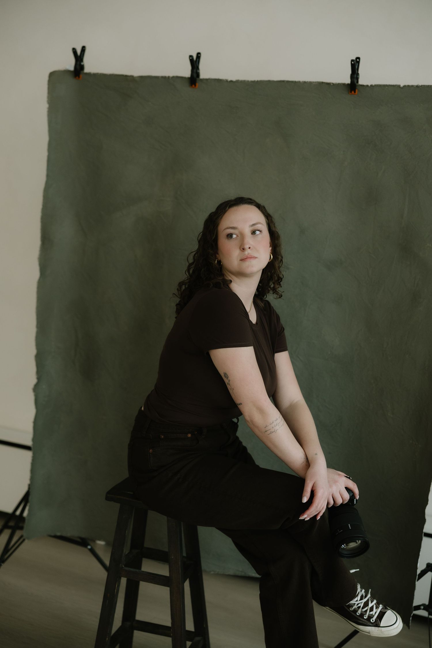 Woman in black outfit sitting on stool against dark green backdrop in photography studio setting.