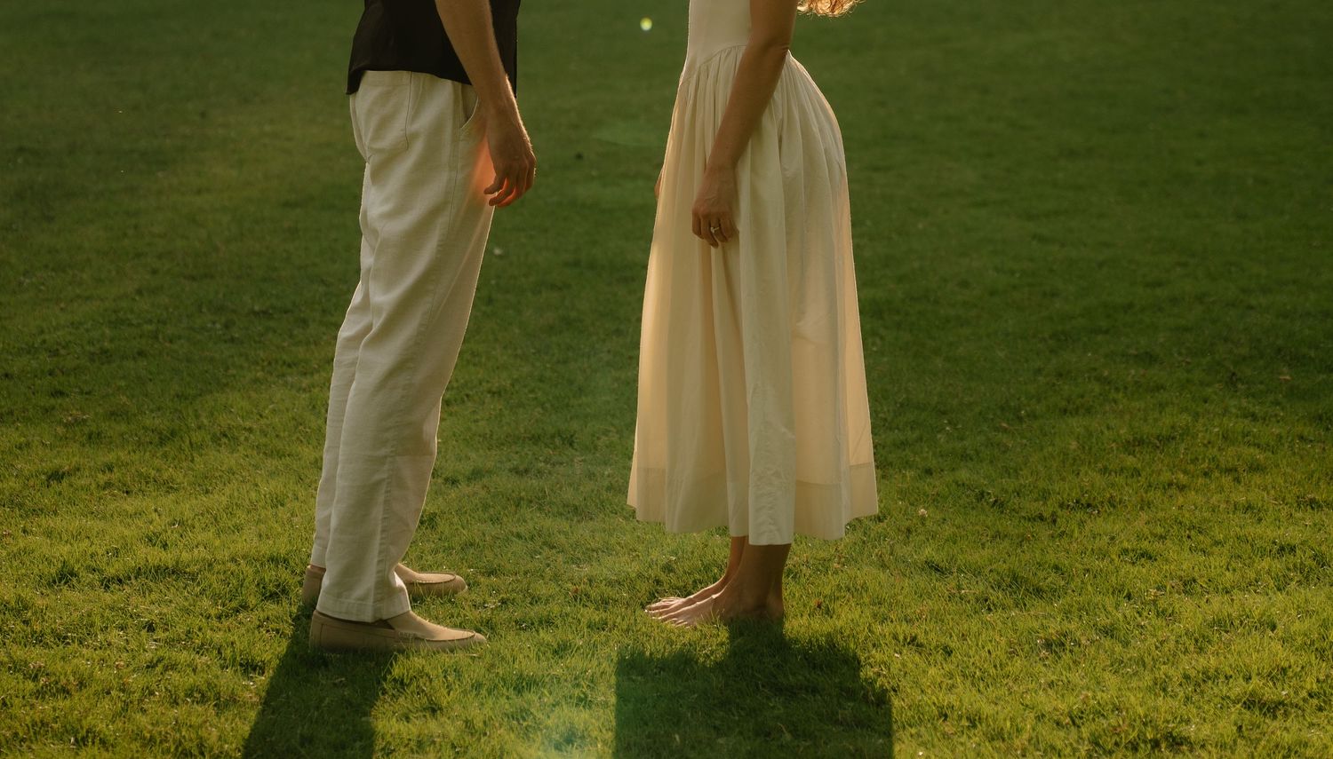 Couple standing barefoot facing each other on a lush green lawn in soft golden evening light.
