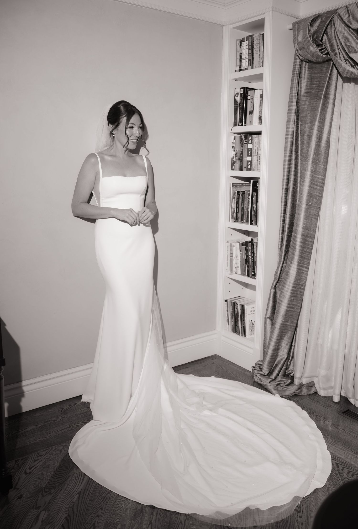 Bride in elegant white satin wedding gown with train poses in black and white photo beside bookshelf and draped curtain.