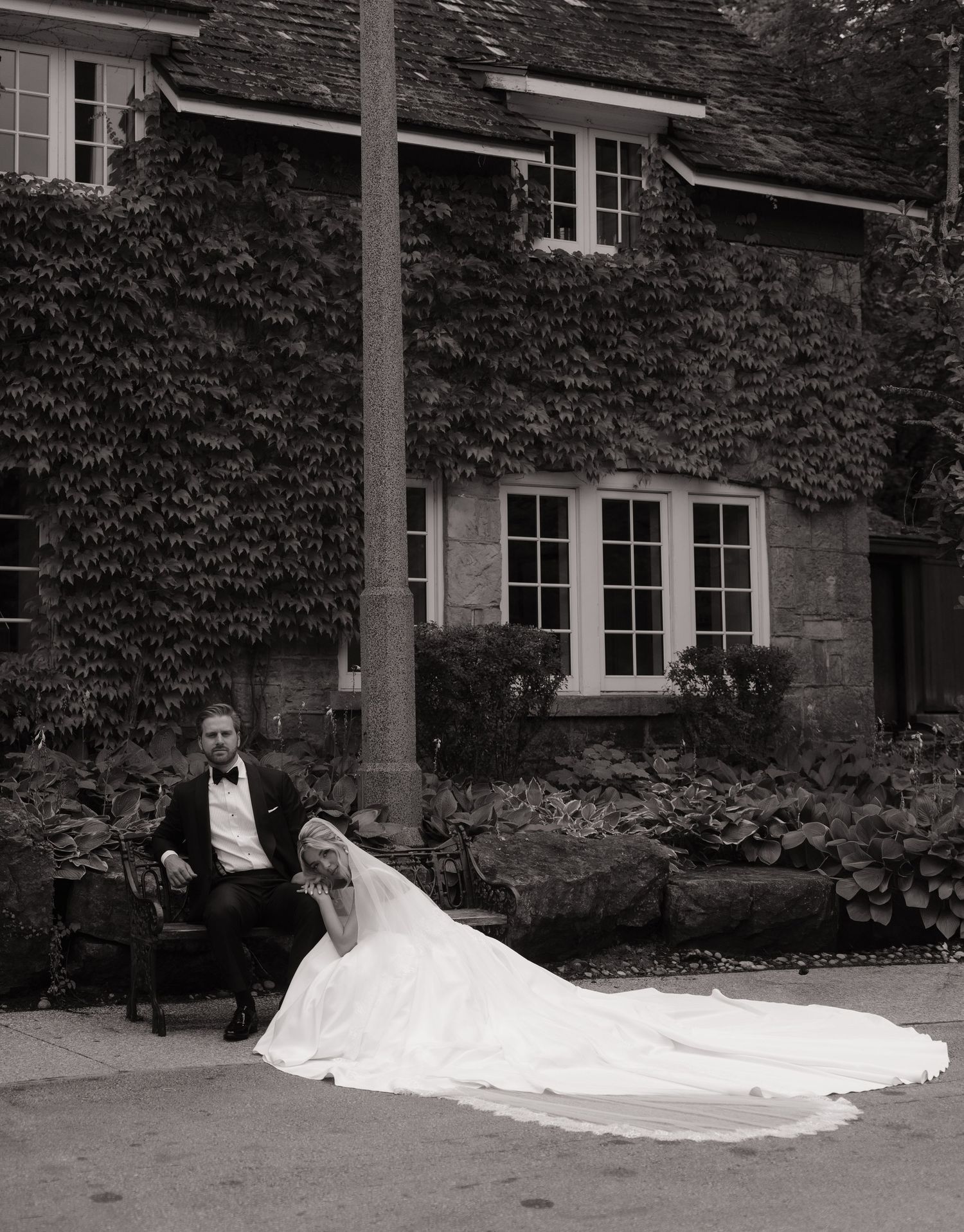 Black and white wedding photo of bride and groom posing outside ivy-covered stone manor house.