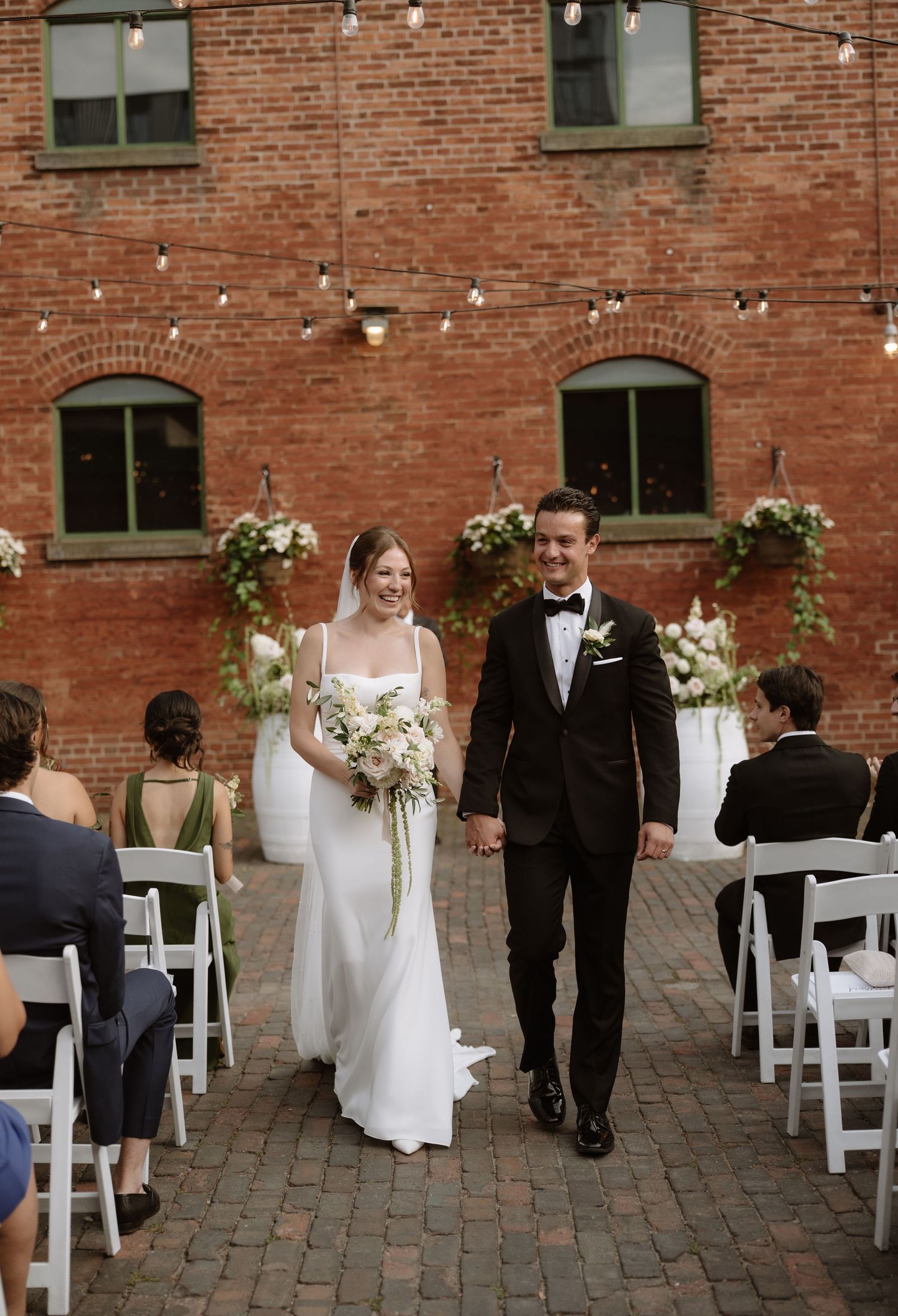 Bride and groom walk down the aisle at an outdoor brick venue wedding ceremony with floral arch backdrop.