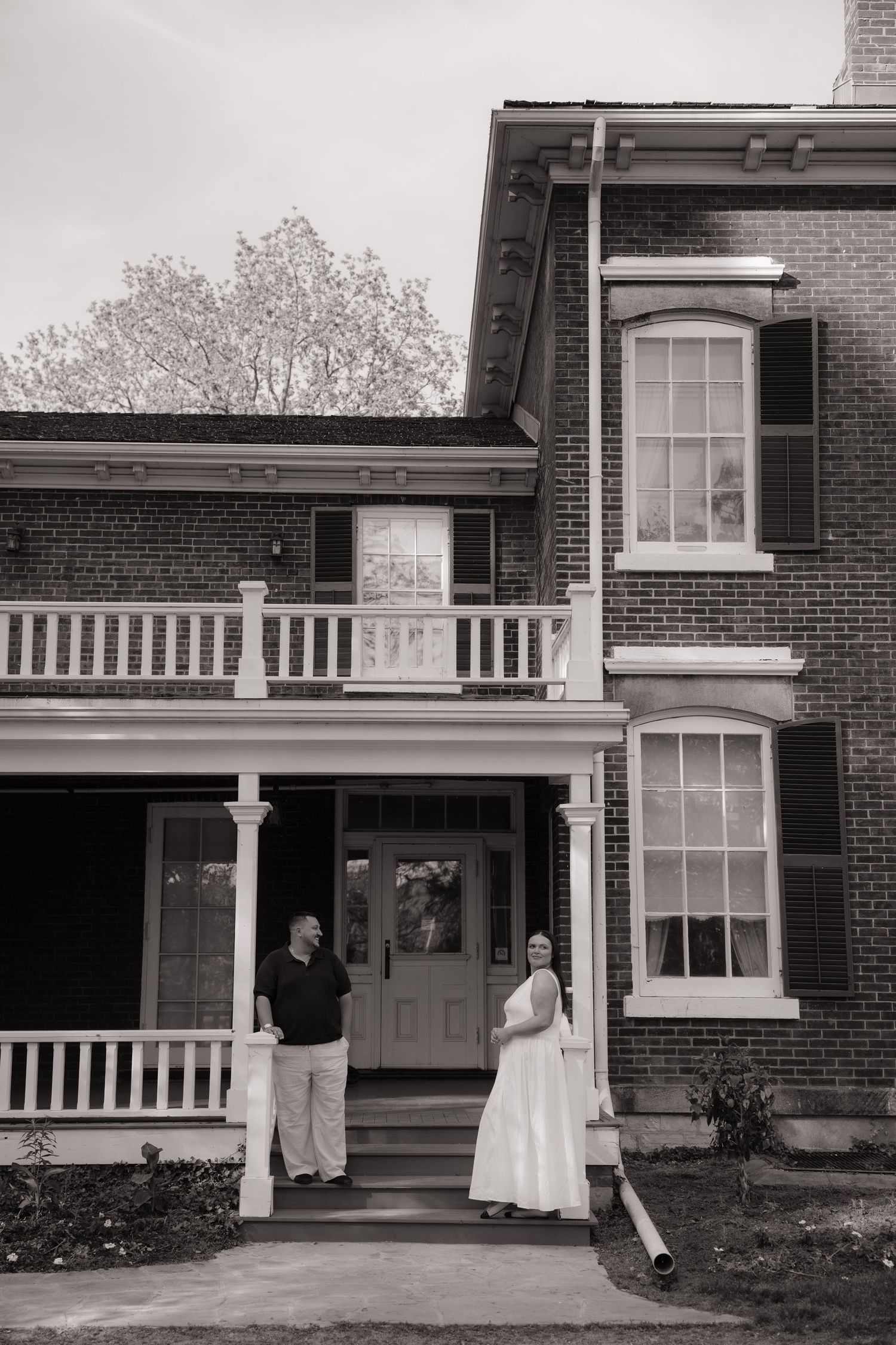 Black and white photo of a couple standing on the porch steps of a classic brick colonial home.