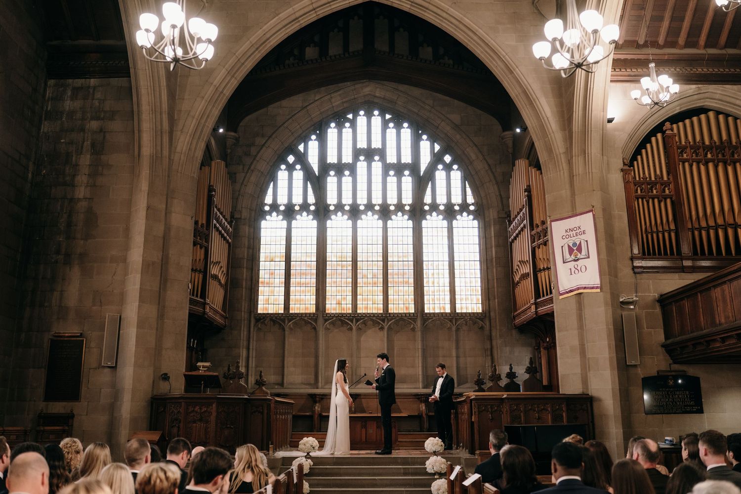 Bride and groom exchange vows at church altar beneath stunning Gothic stained glass window before seated guests.