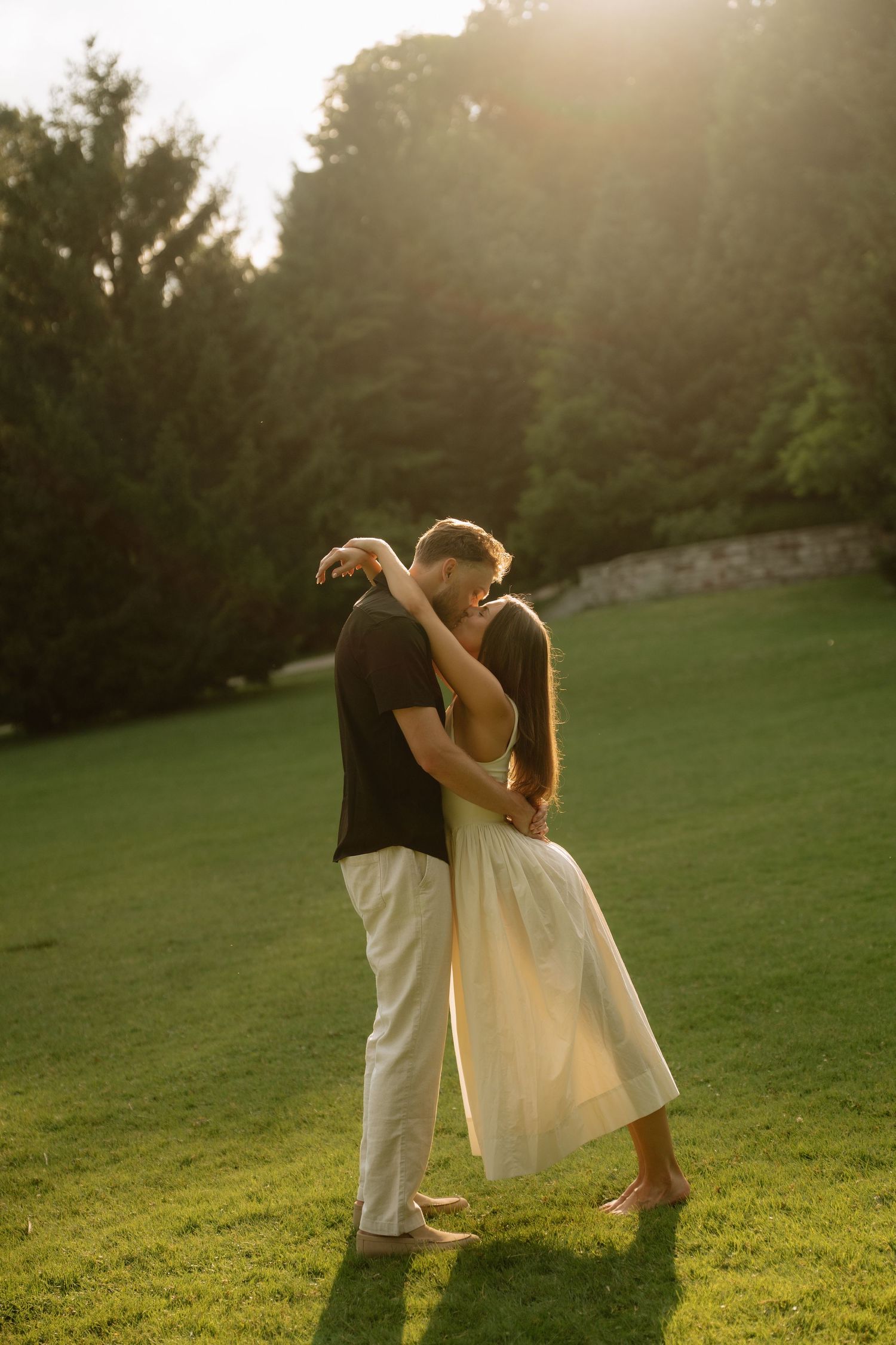 Couple embracing and kissing on green lawn at golden hour sunset with trees in background.