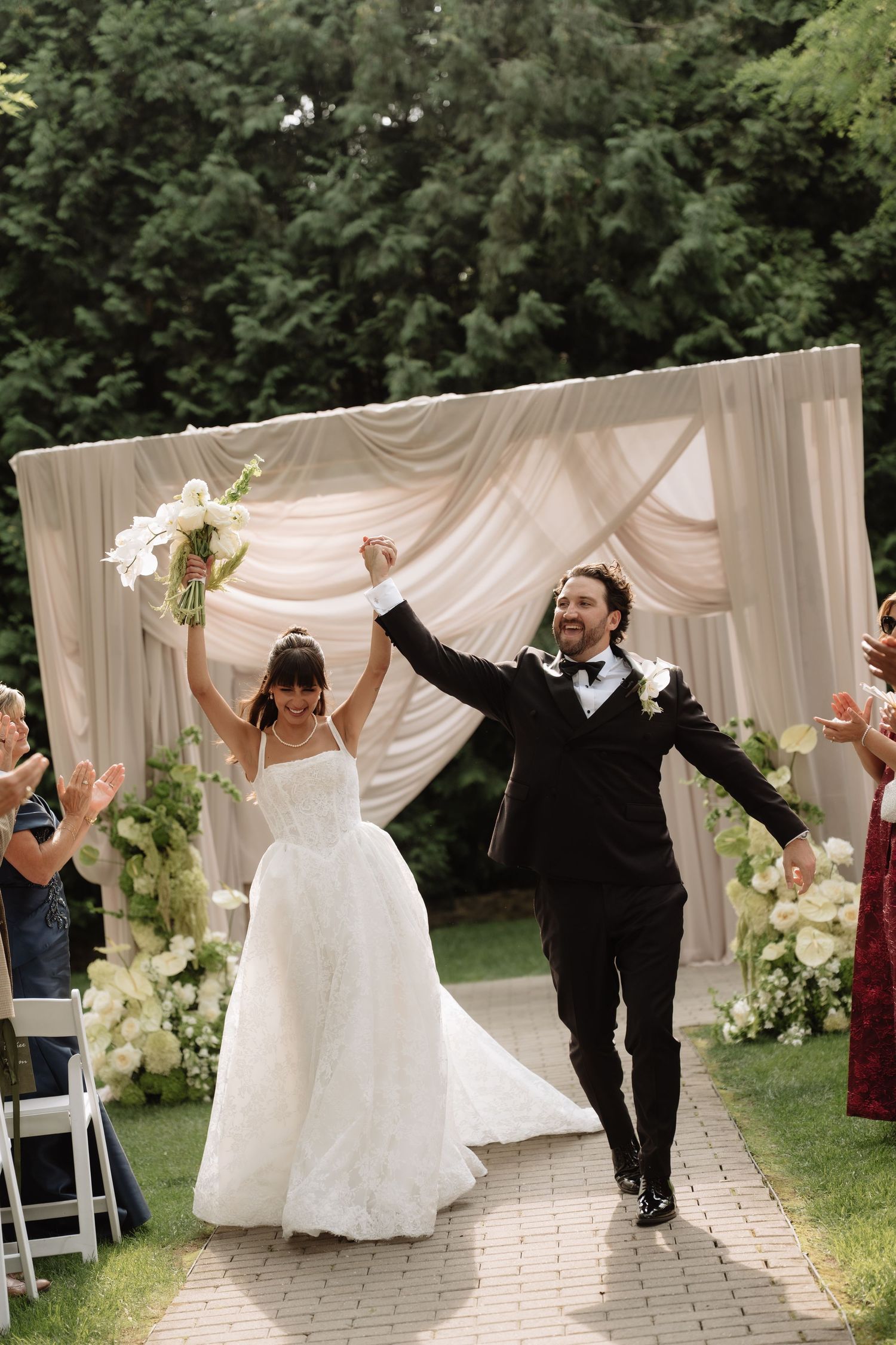 Joyful newlyweds celebrate recessional at outdoor wedding ceremony with floral arch and white draping.