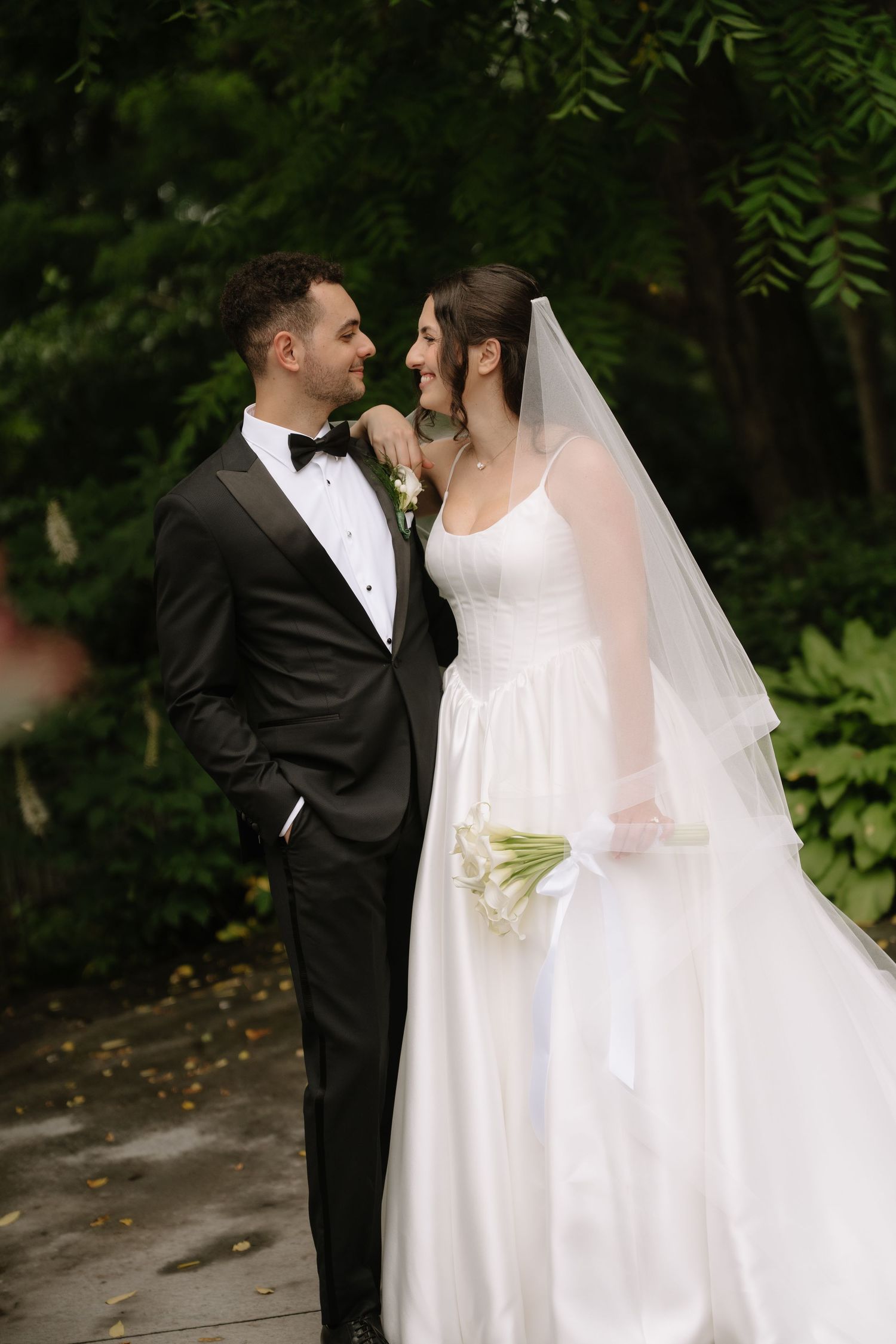 Bride and groom gazing at each other in a lush green garden, bride holding white calla lily bouquet.