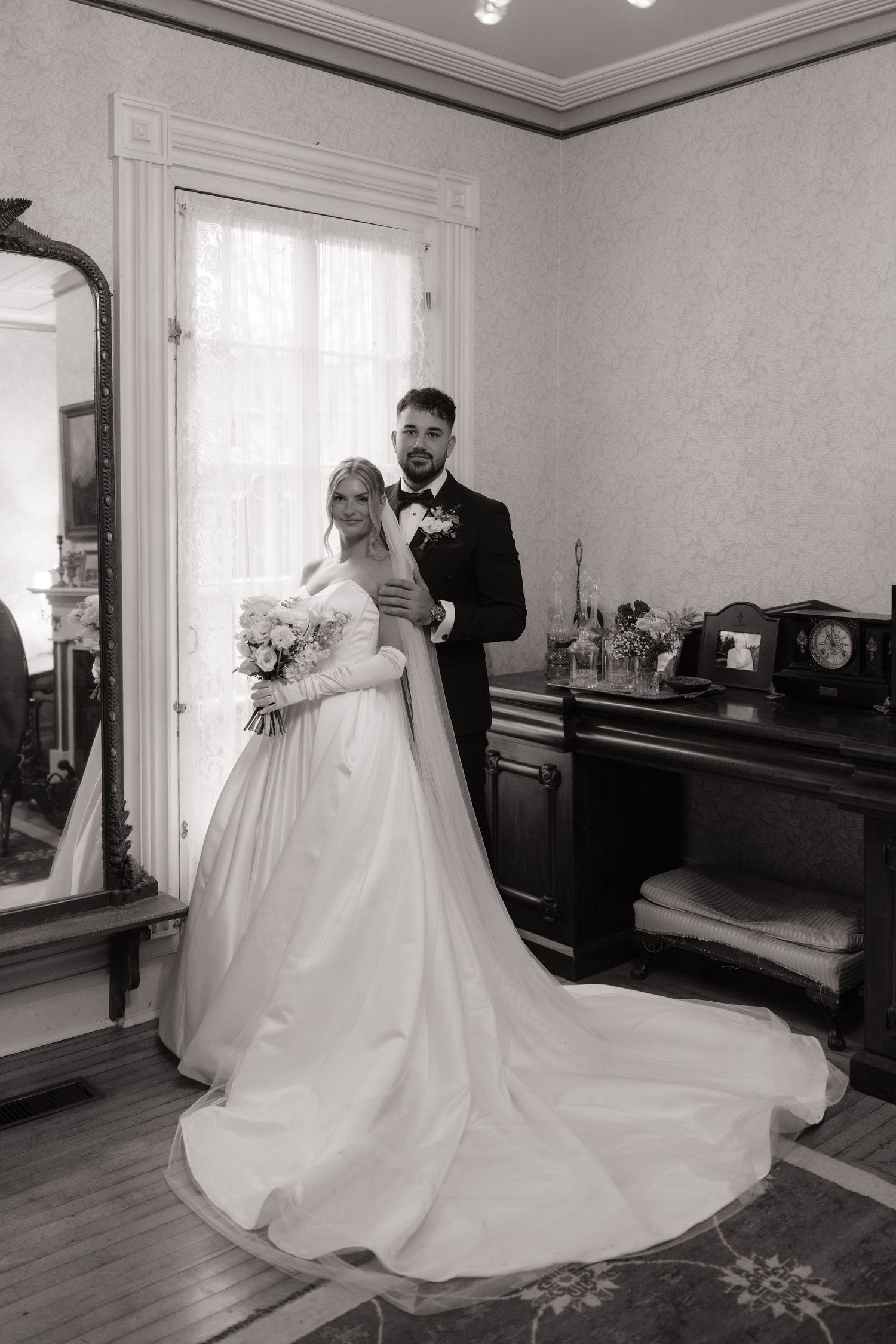 Black and white portrait of bride in ballgown and groom embracing in an elegant vintage-styled room.
