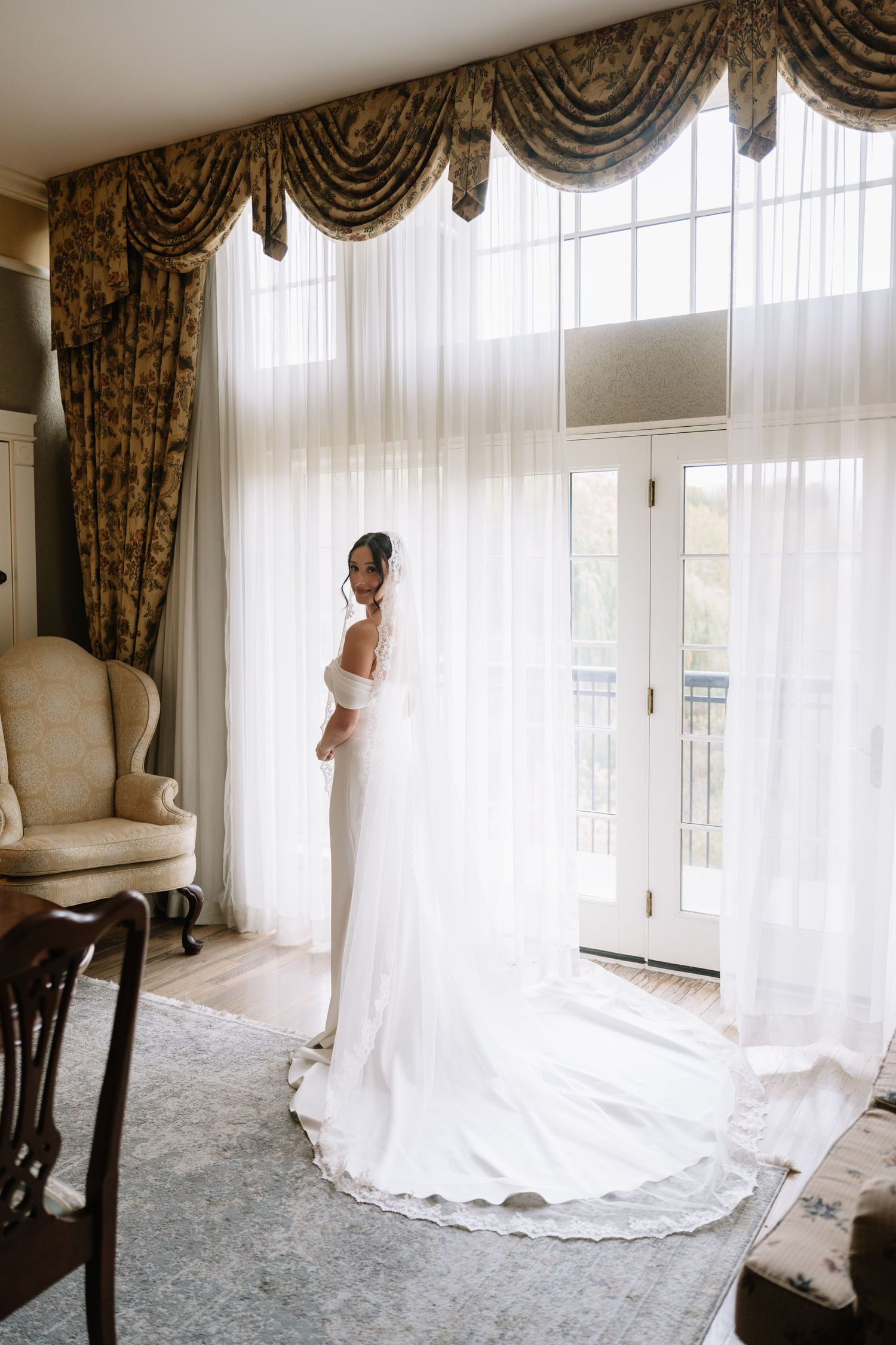 Elegant bride in off-shoulder wedding gown with cathedral veil standing by sheer curtained windows.