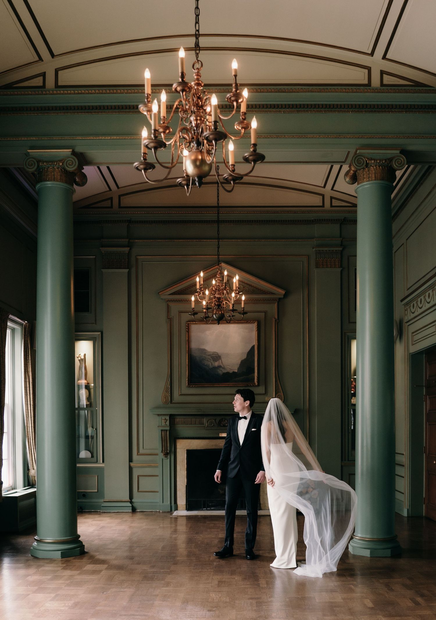 Bride and groom pose in elegant green ballroom with chandelier and tall columns.