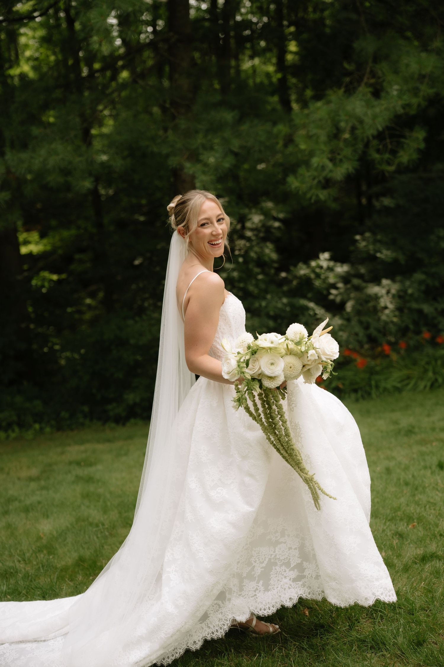 Smiling bride in white lace gown and veil holds white bouquet outdoors in lush green garden setting.