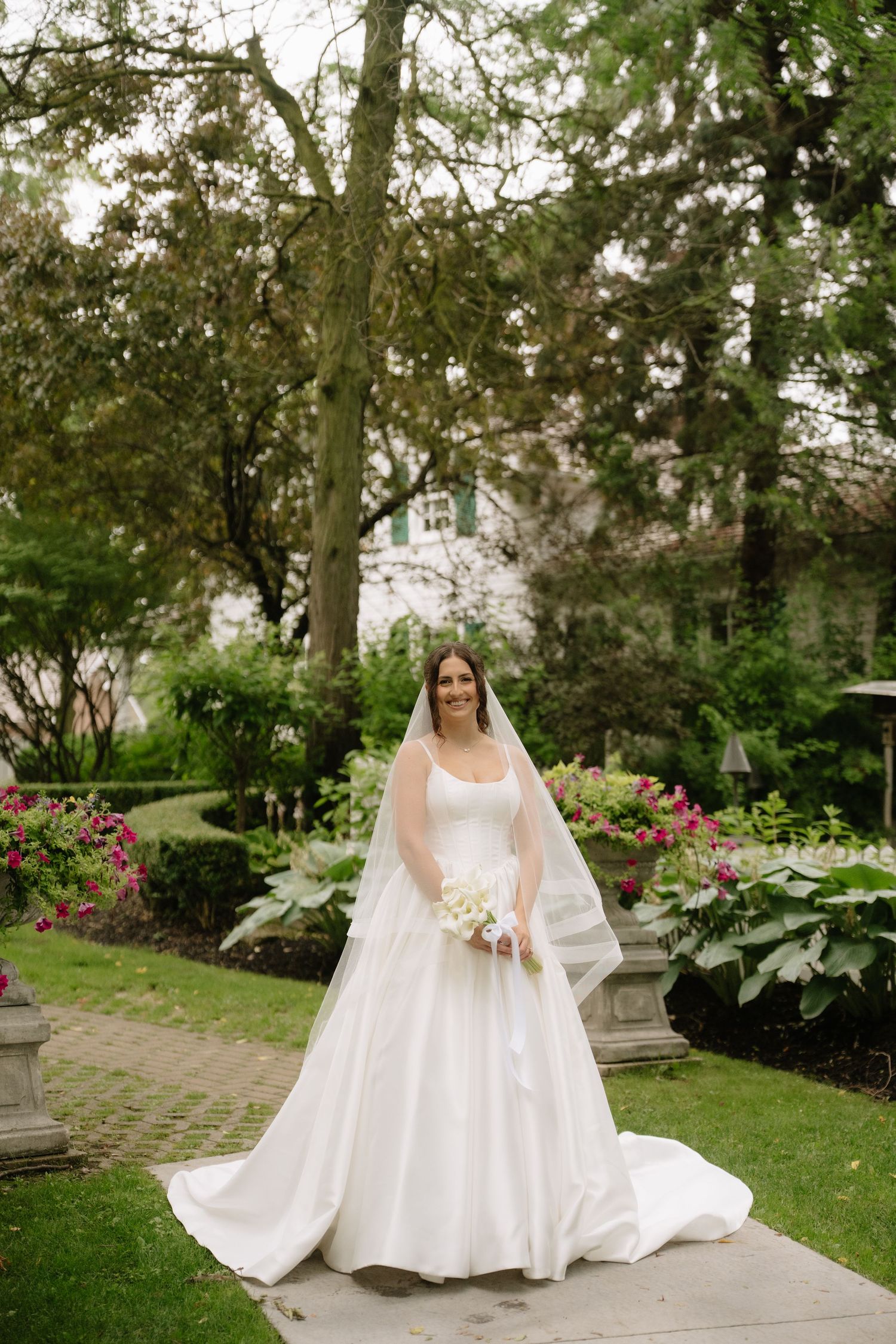 Smiling bride in a white ball gown and veil holding a bouquet in a beautiful garden setting.