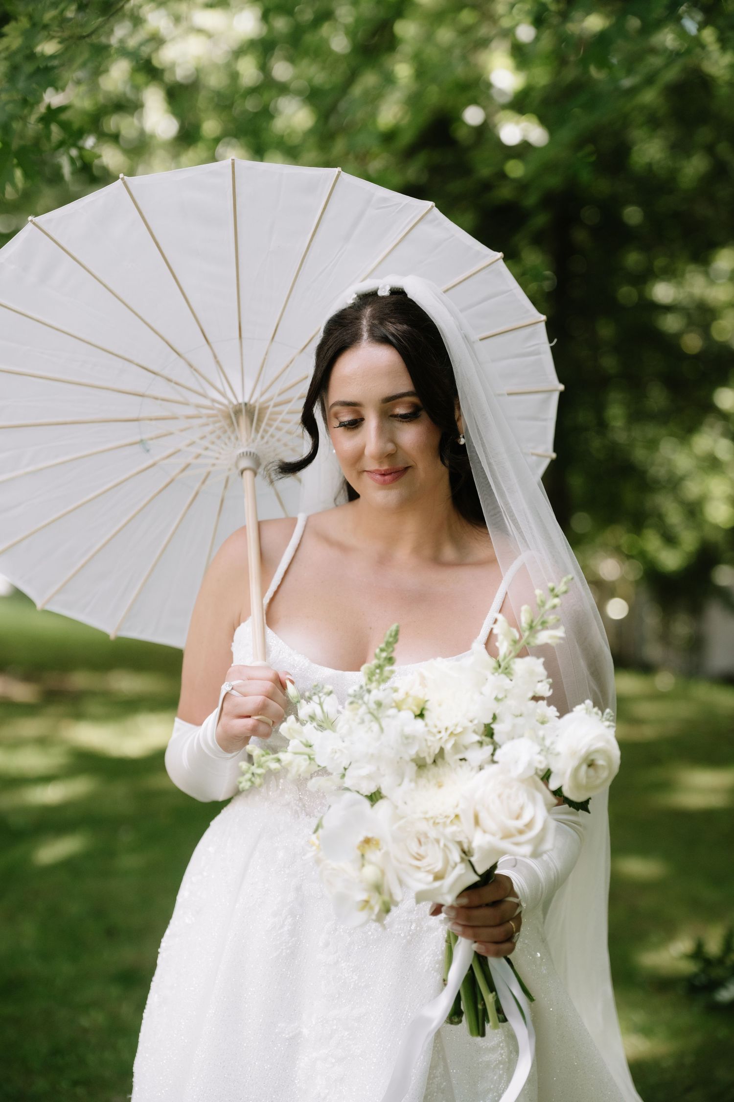 Bride in white lace gown holds white floral bouquet and parasol umbrella outdoors in green garden setting.