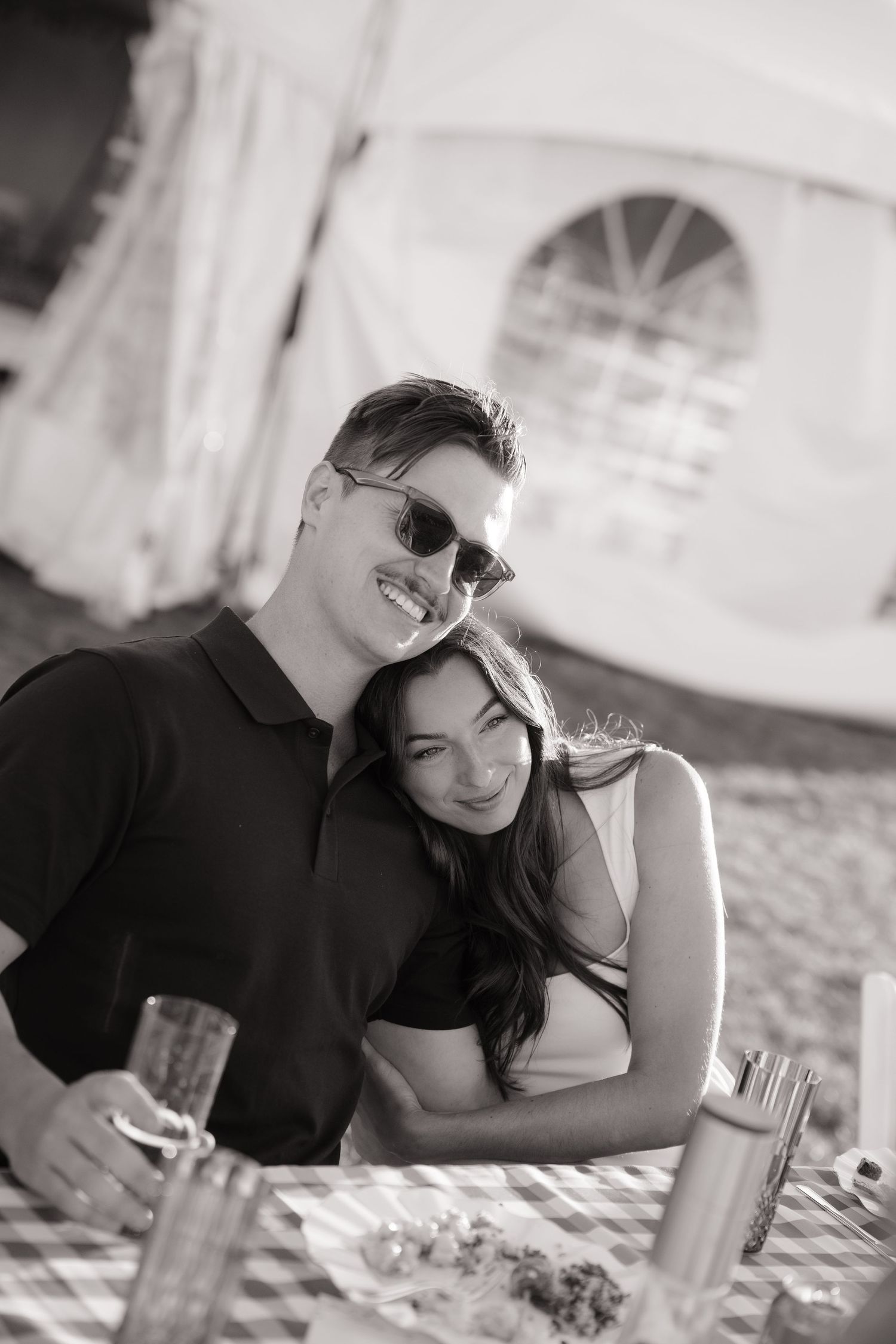 Black and white photo of smiling couple embracing at outdoor rehearsal dinner table with tent in background.