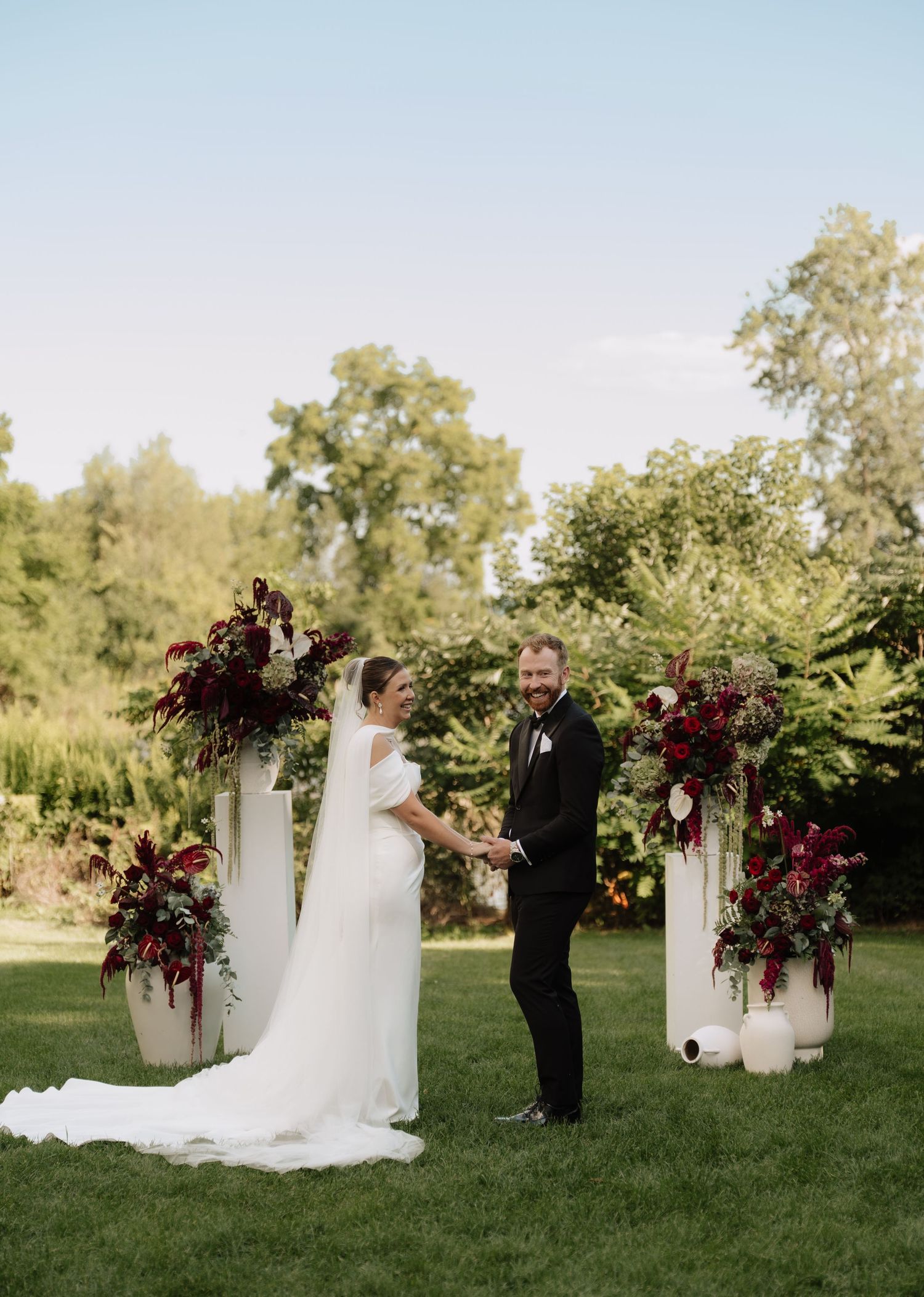 Bride and groom exchange vows outdoors flanked by tall white pedestals with deep red floral arrangements.