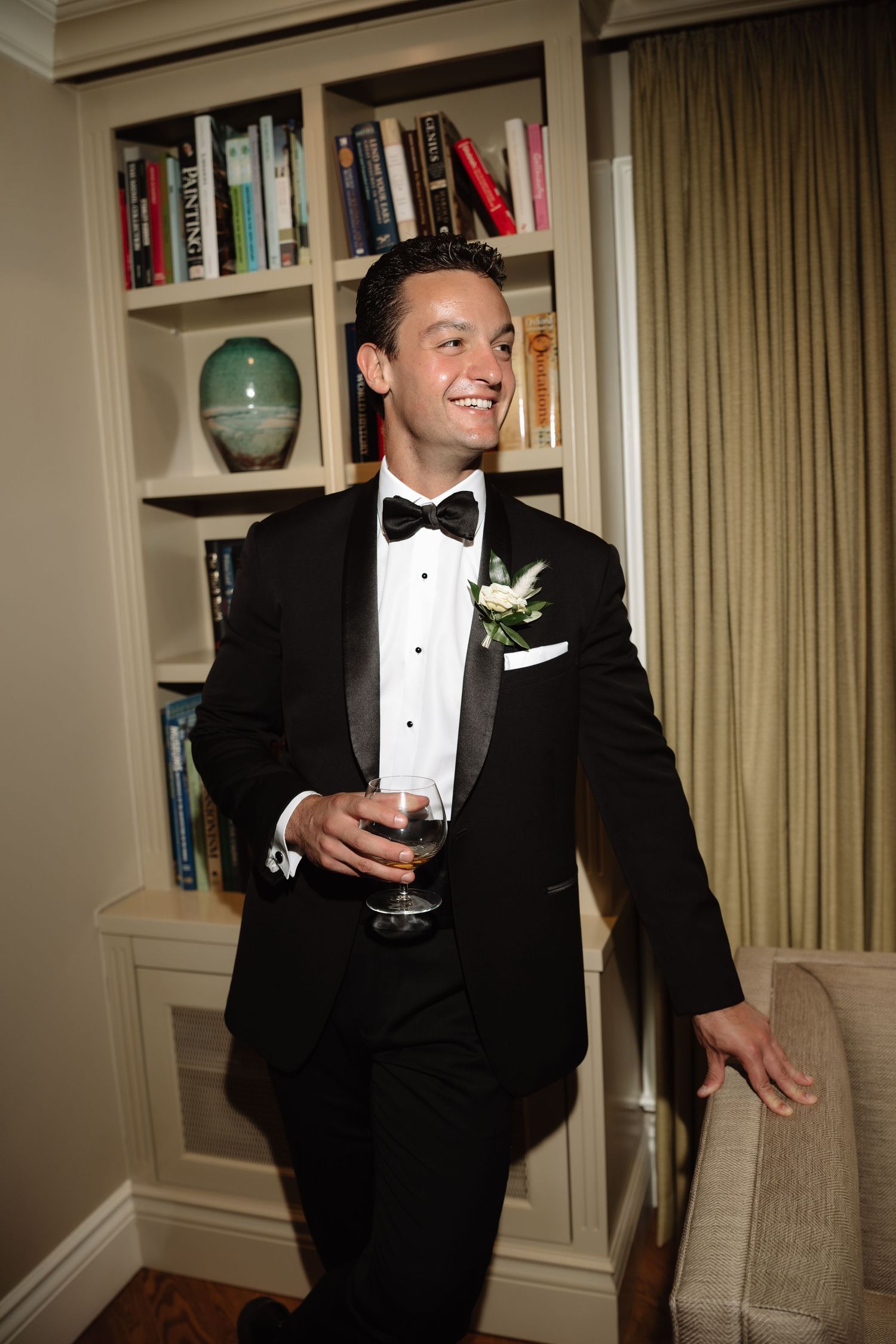 Smiling groom in black tuxedo with bow tie and boutonniere holds champagne glass near bookshelf.
