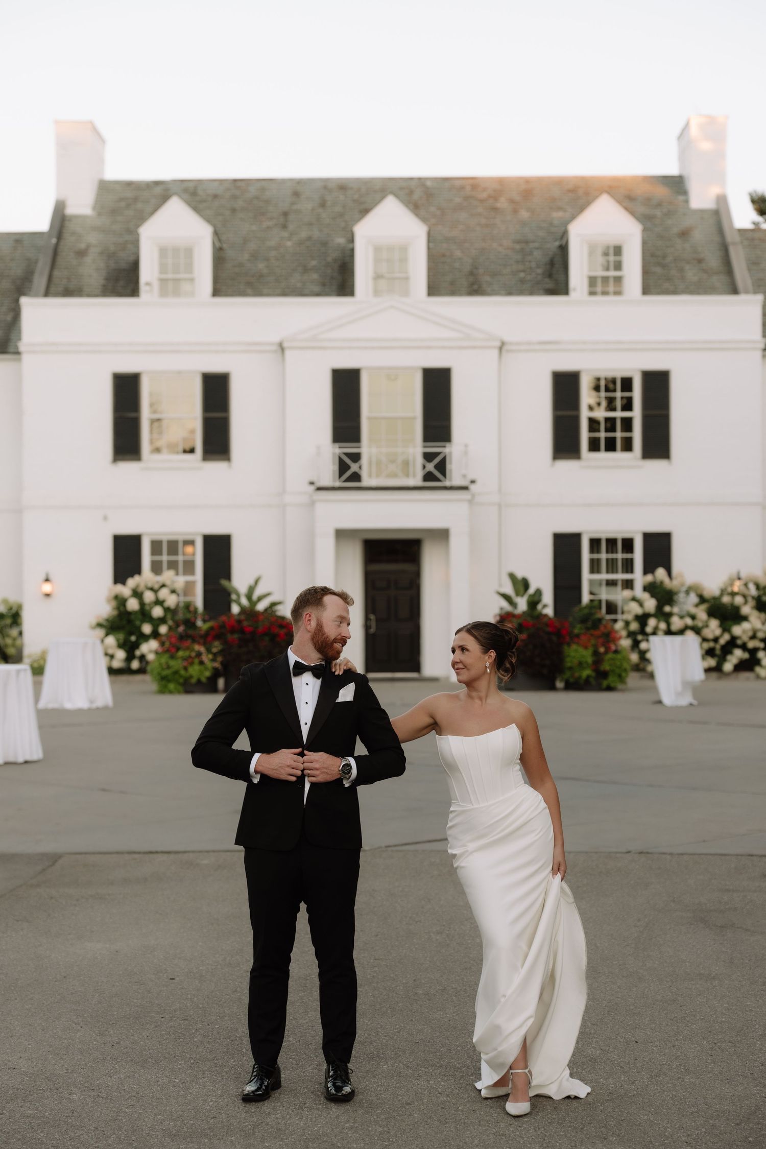 Bride and groom share a joyful moment outside a elegant white colonial mansion with black shutters.