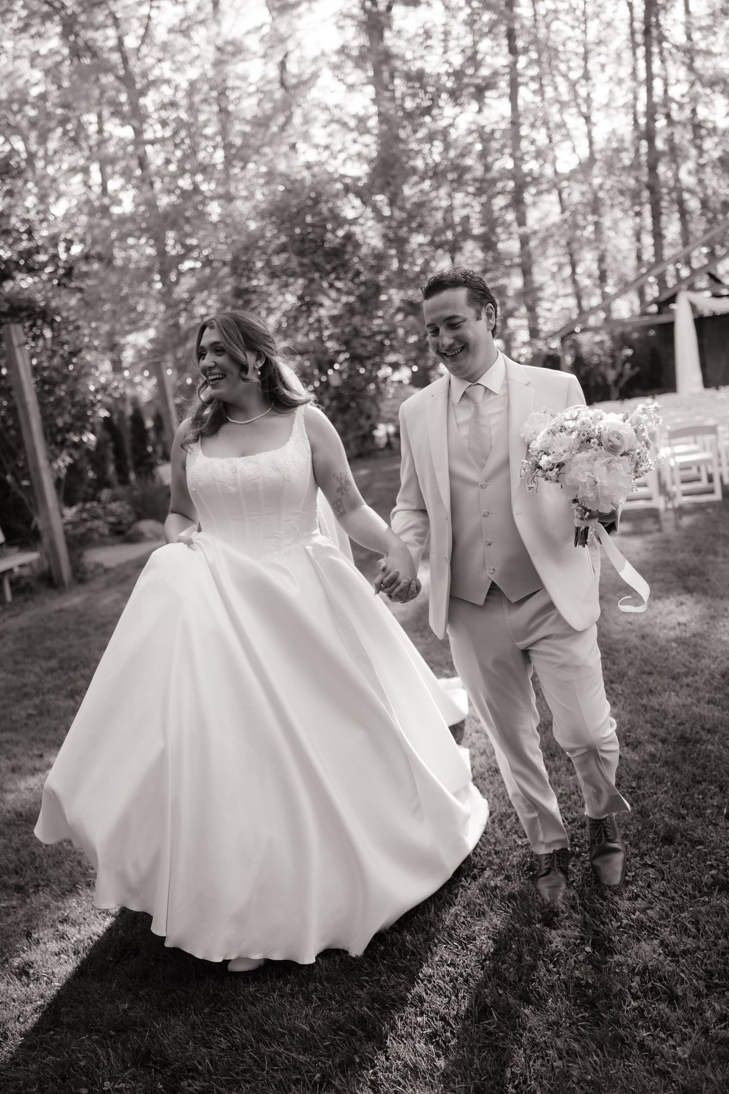 Black and white photo of smiling bride and groom holding hands walking through outdoor garden venue.