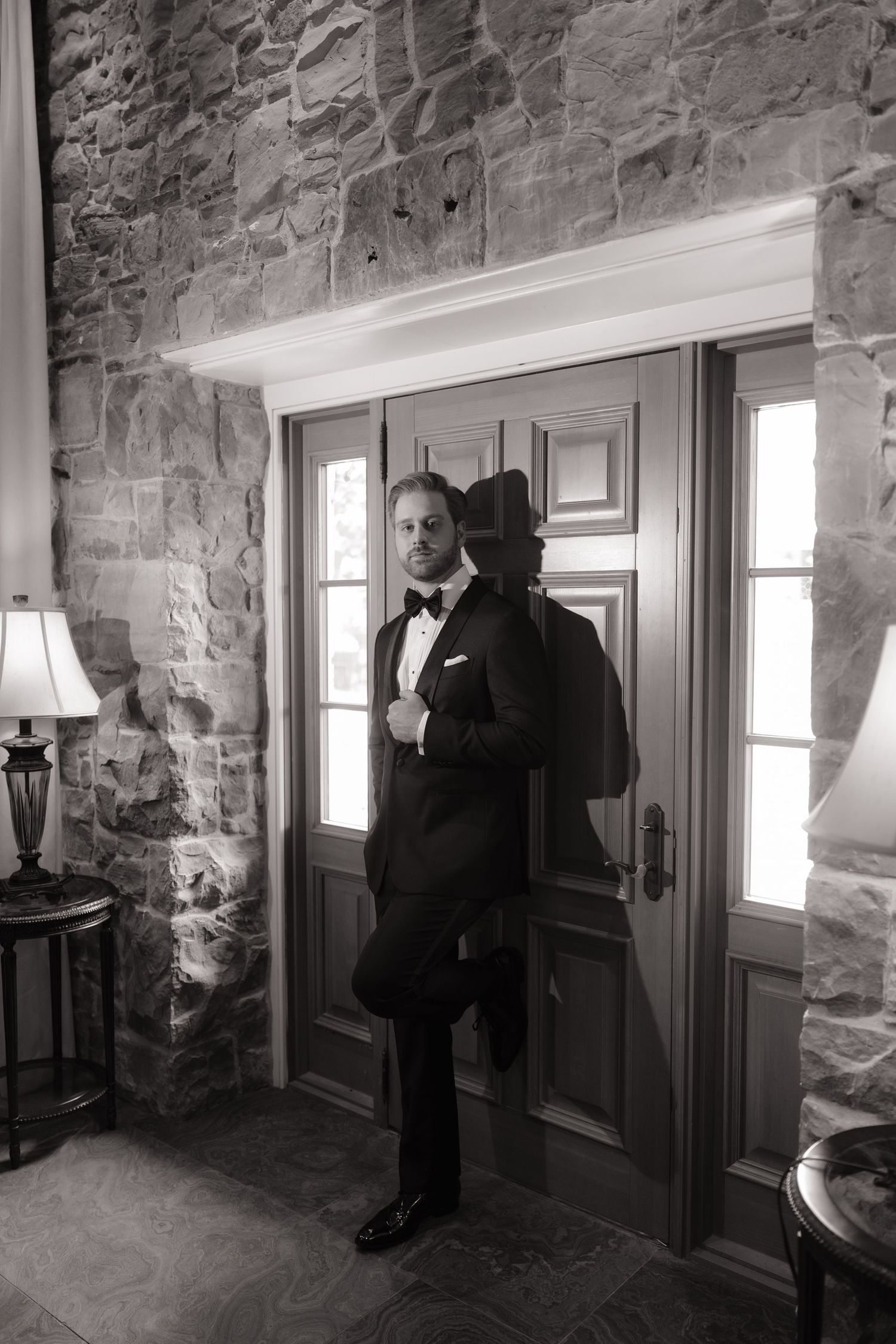 Groom in black tuxedo and bow tie poses by stone wall entrance in elegant black and white wedding portrait.
