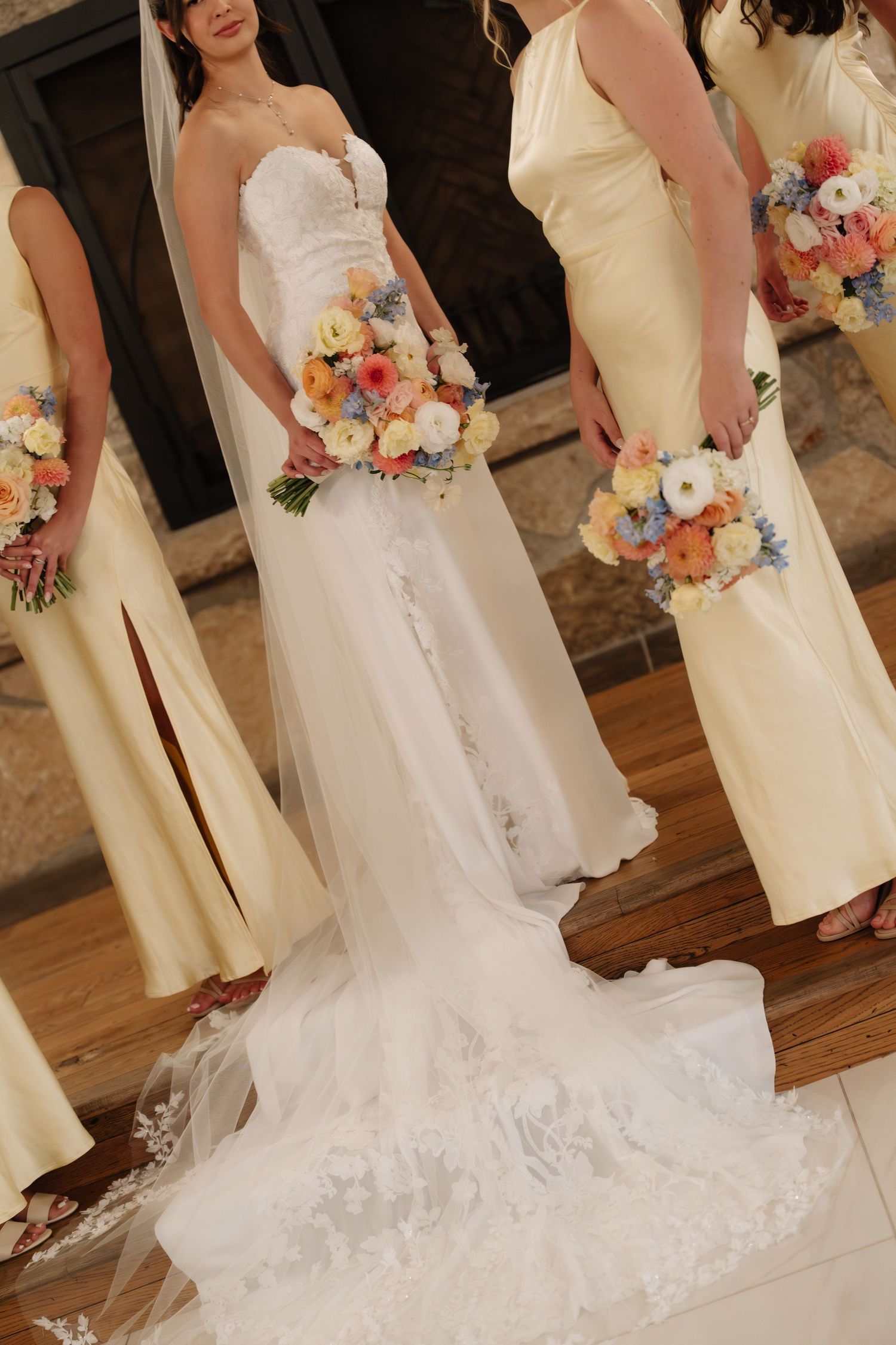 Bride in lace wedding gown with bridesmaids in yellow dresses holding colorful bouquets indoors.