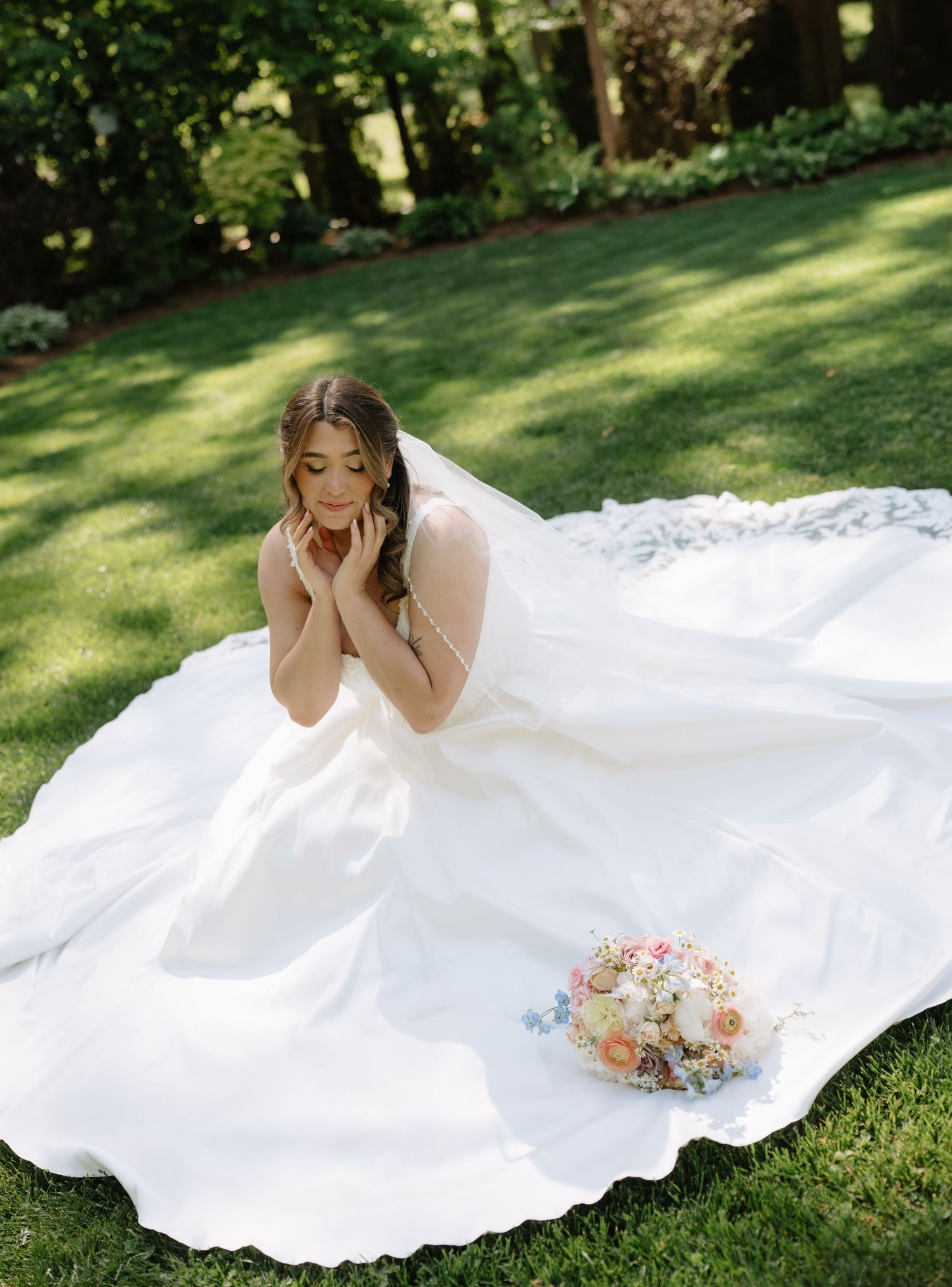 Bride in stunning white ball gown sitting on lush green grass with flowing train and bridal bouquet.