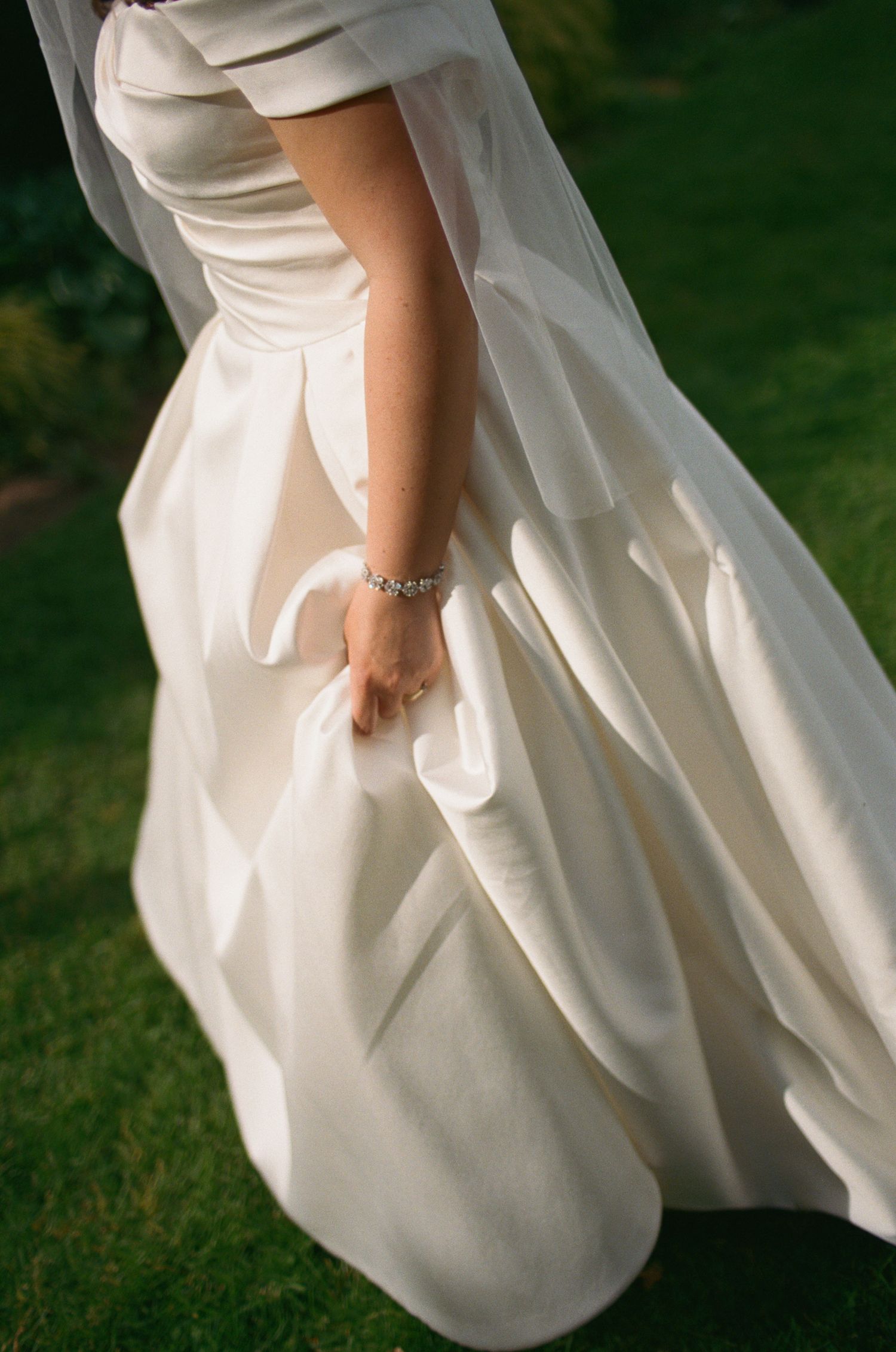 Close-up of bride holding voluminous ivory satin wedding gown skirt, showcasing delicate diamond bracelet on wrist.