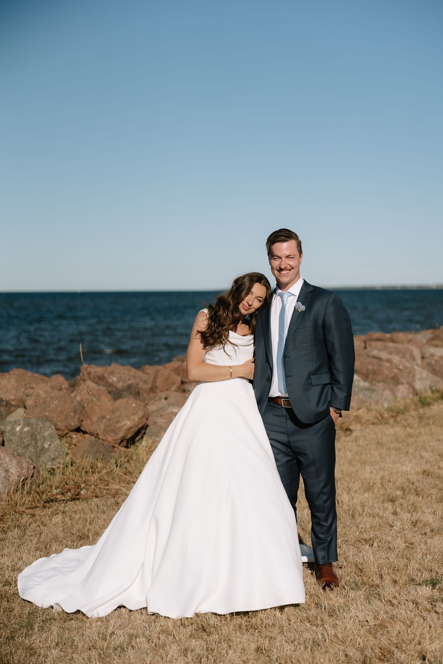 Bride and groom posing together on rocky shoreline in white gown and navy suit with ocean backdrop.