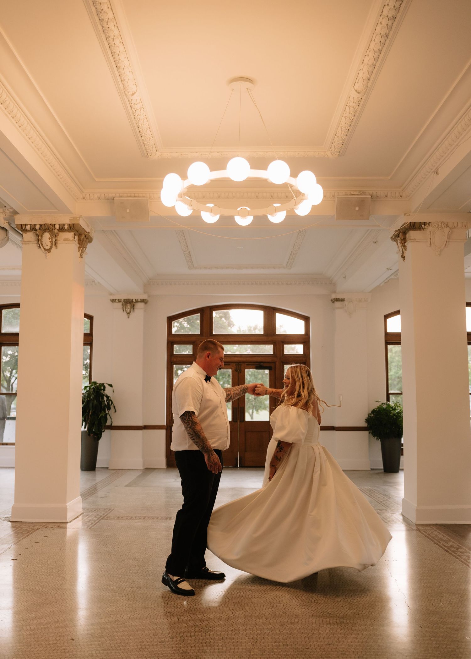 Couple sharing first dance in elegant white venue with ornate ceiling and chandelier.
