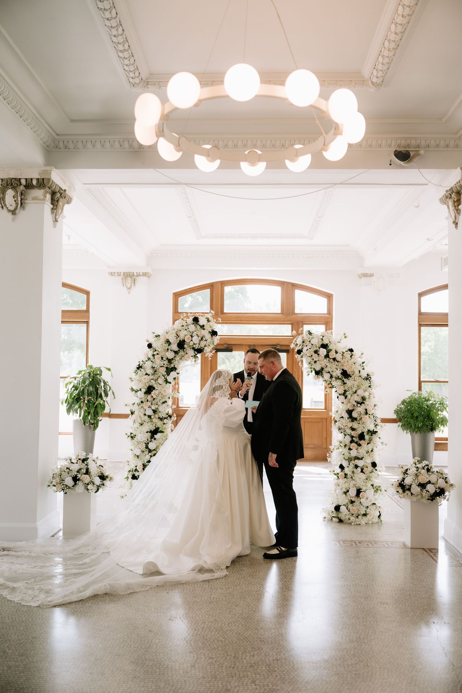 Bride and groom stand under white floral arch in elegant white ceremony hall with ornate ceiling details.