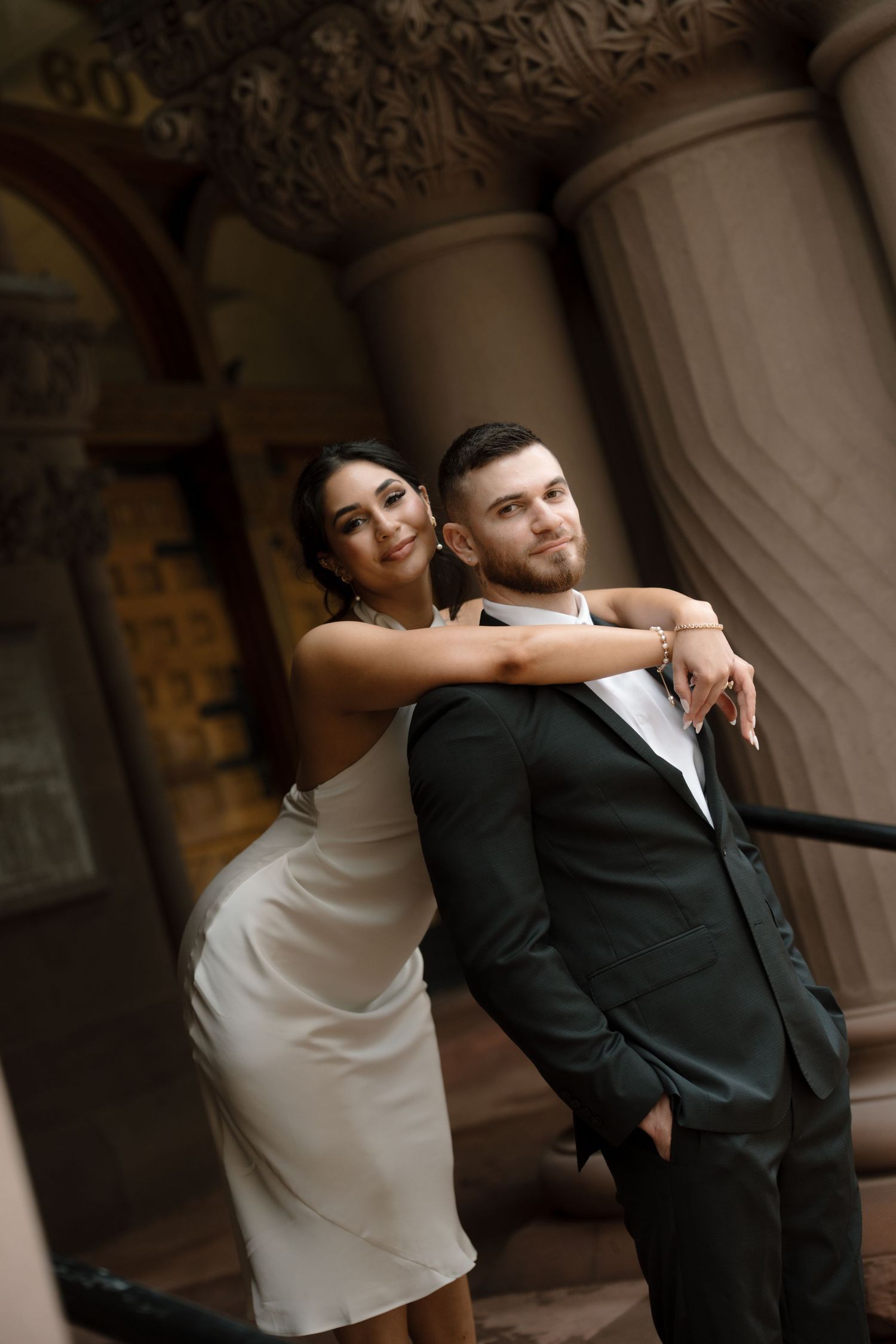 Bride in white dress embracing groom in dark suit beside decorative stone columns of historic venue.