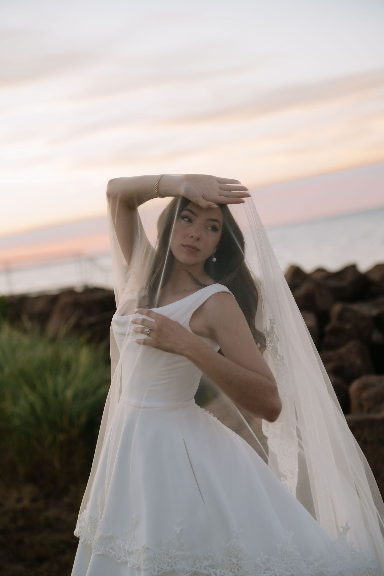 Elegant bride posing with veil overhead in white gown at sunset near rocky ocean shoreline.
