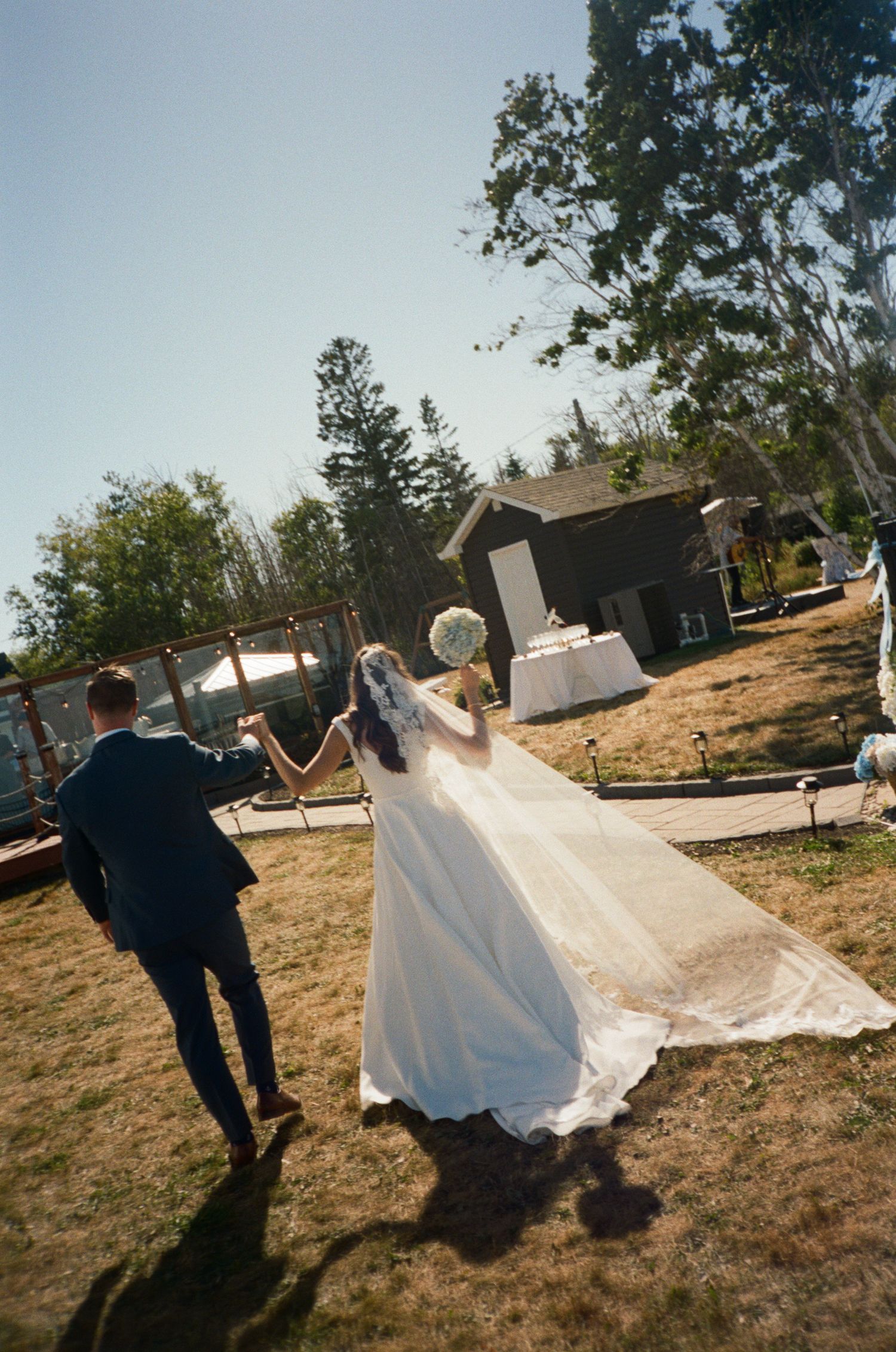 Bride in flowing white gown and groom in navy suit walking hand in hand toward outdoor wedding venue.