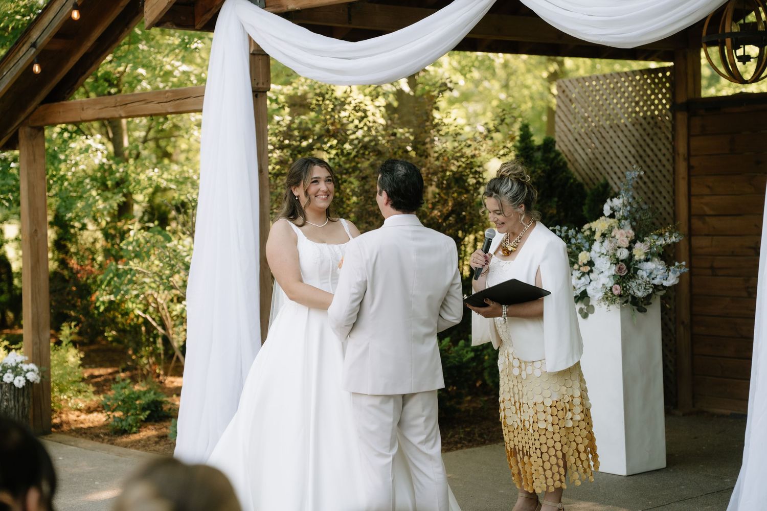 Bride and groom exchanging vows under draped arbor with officiant at outdoor garden wedding ceremony.