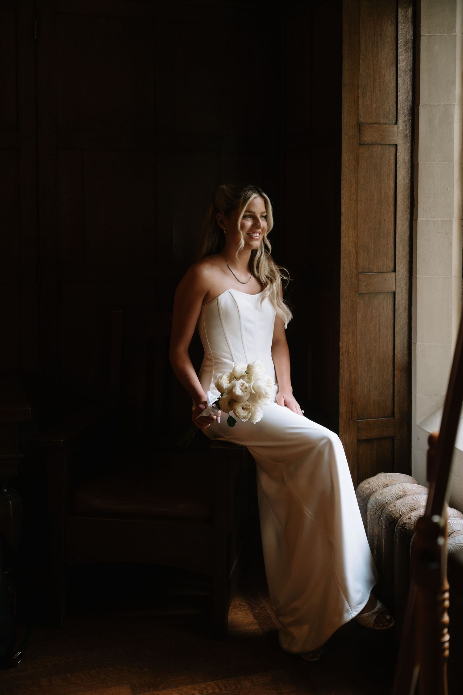 Smiling bride in white sleeveless gown holding ivory bouquet, seated by dark wooden door in moody natural light.