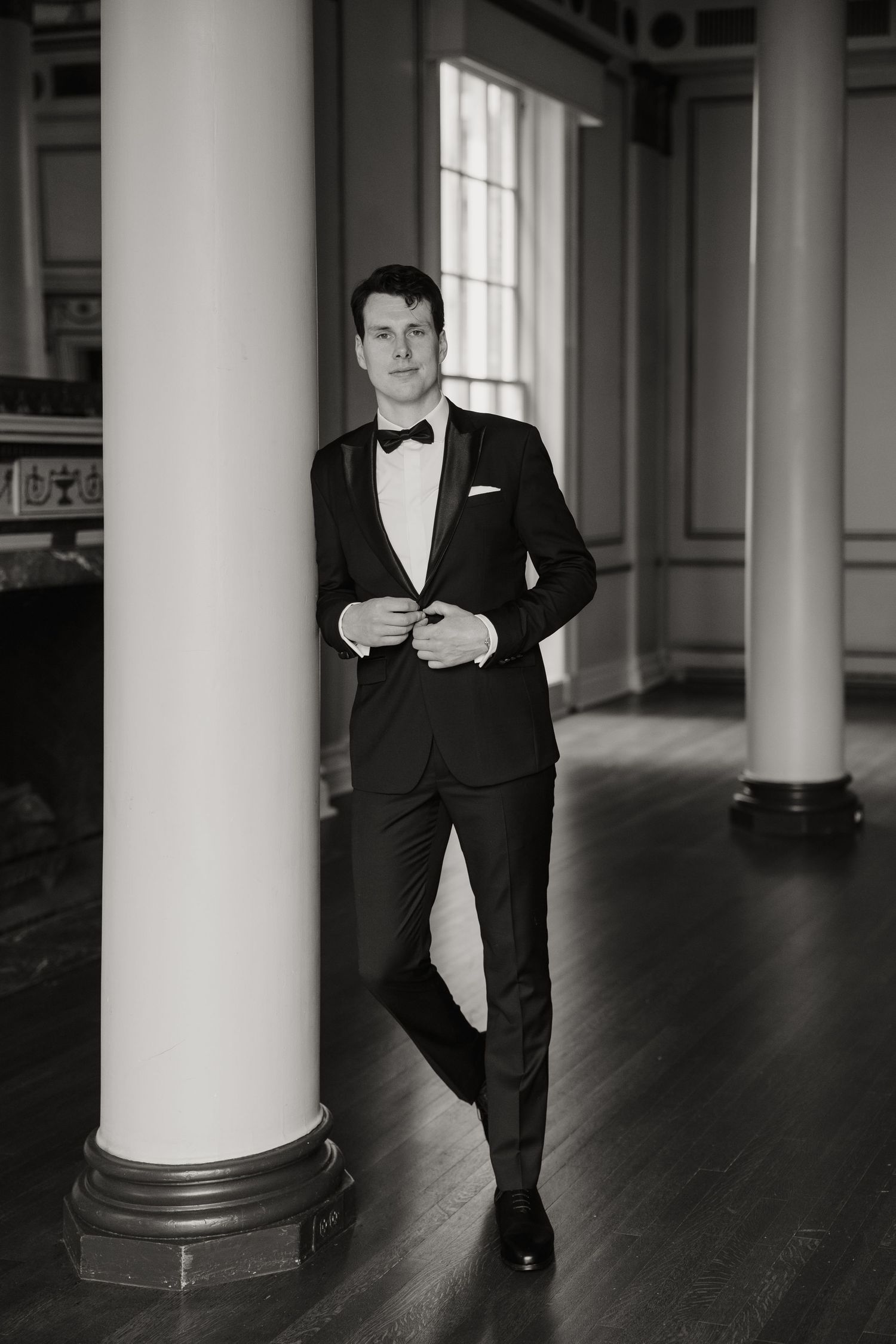 Groom in black tuxedo with bow tie leaning against white column in elegant ballroom, black and white photo.