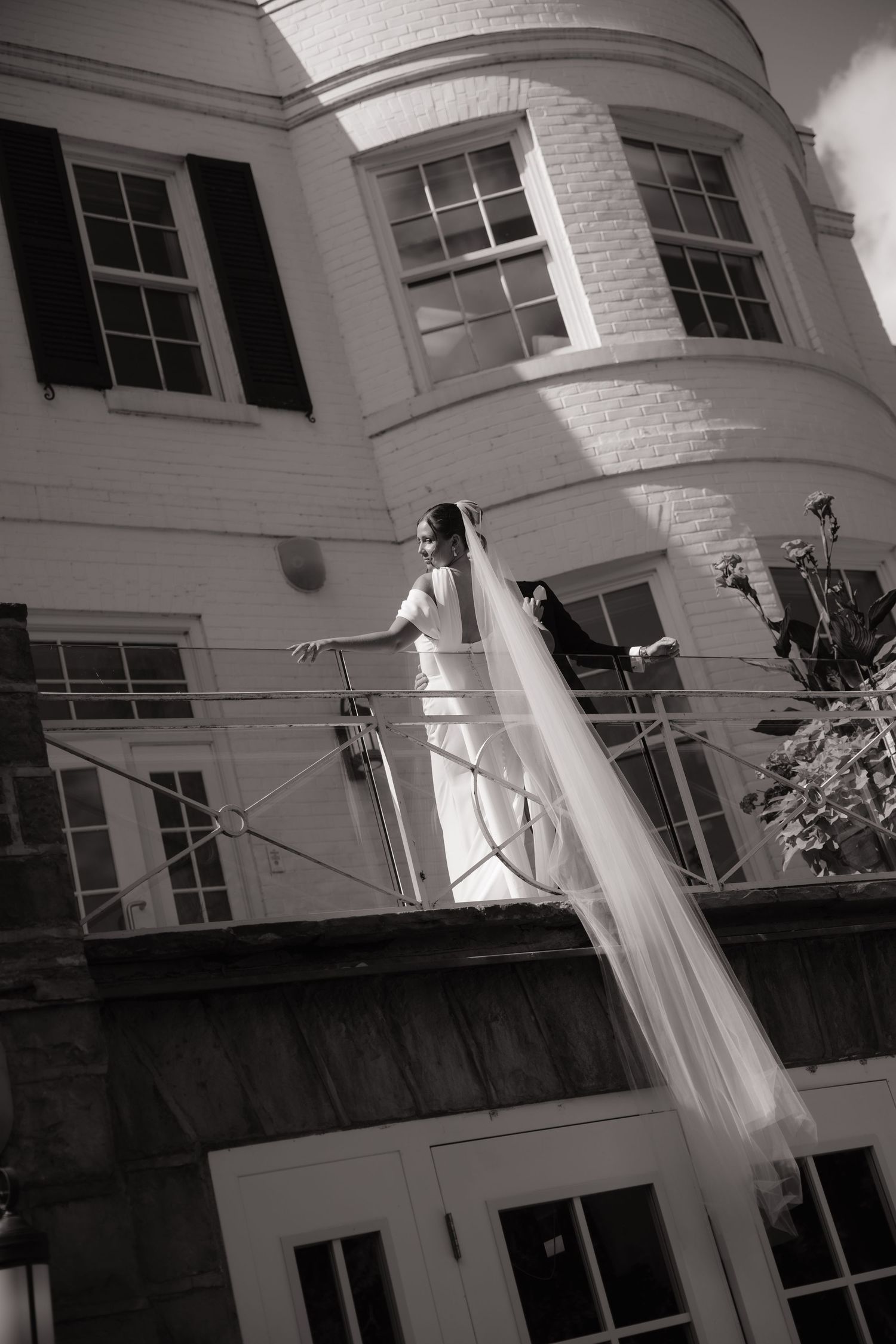 Black and white photo of a bride in a long veil standing on a balcony outside an elegant brick building.