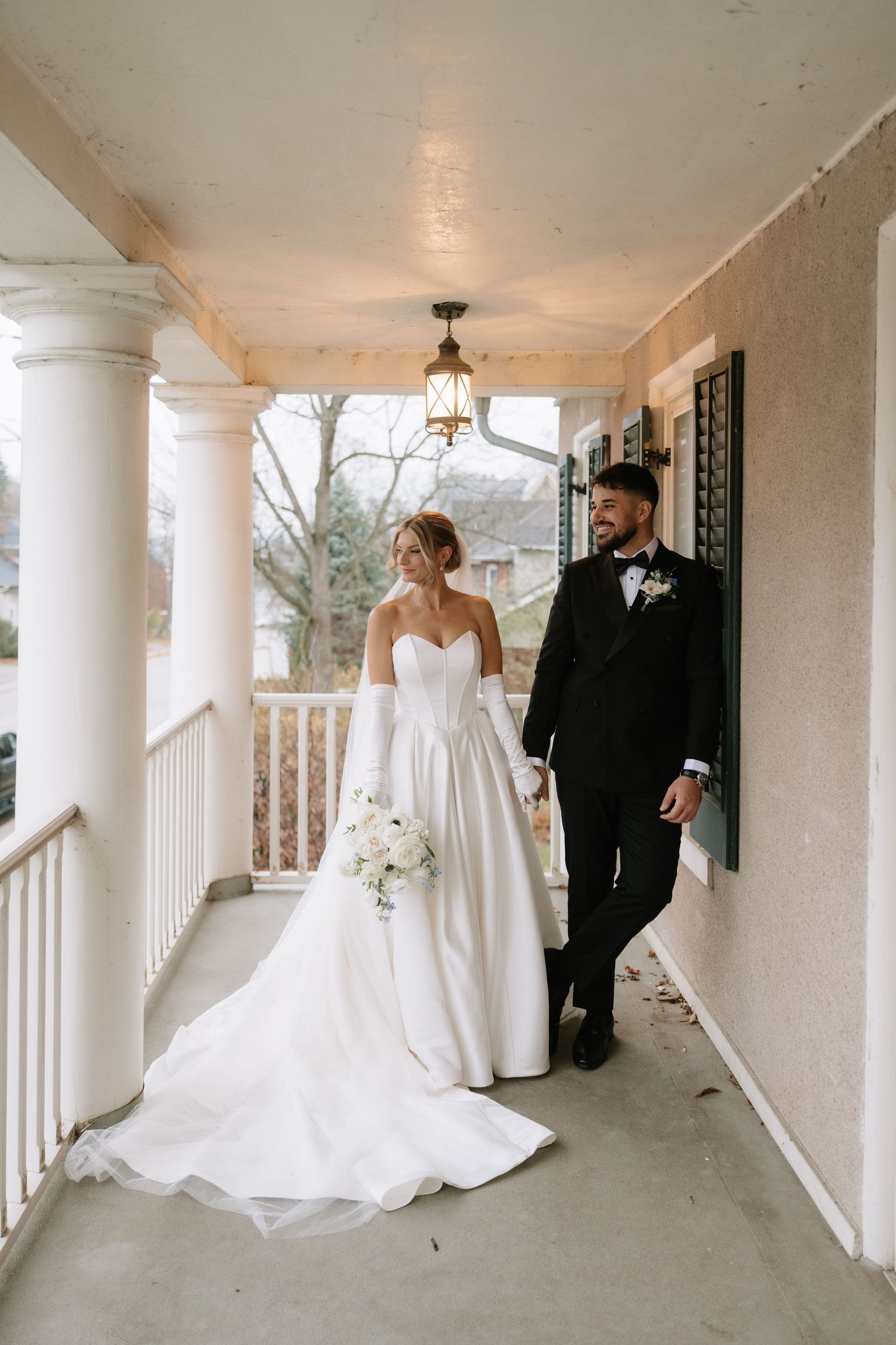 Bride in strapless ballgown with gloves and groom in black tuxedo walking on covered porch of historic home.